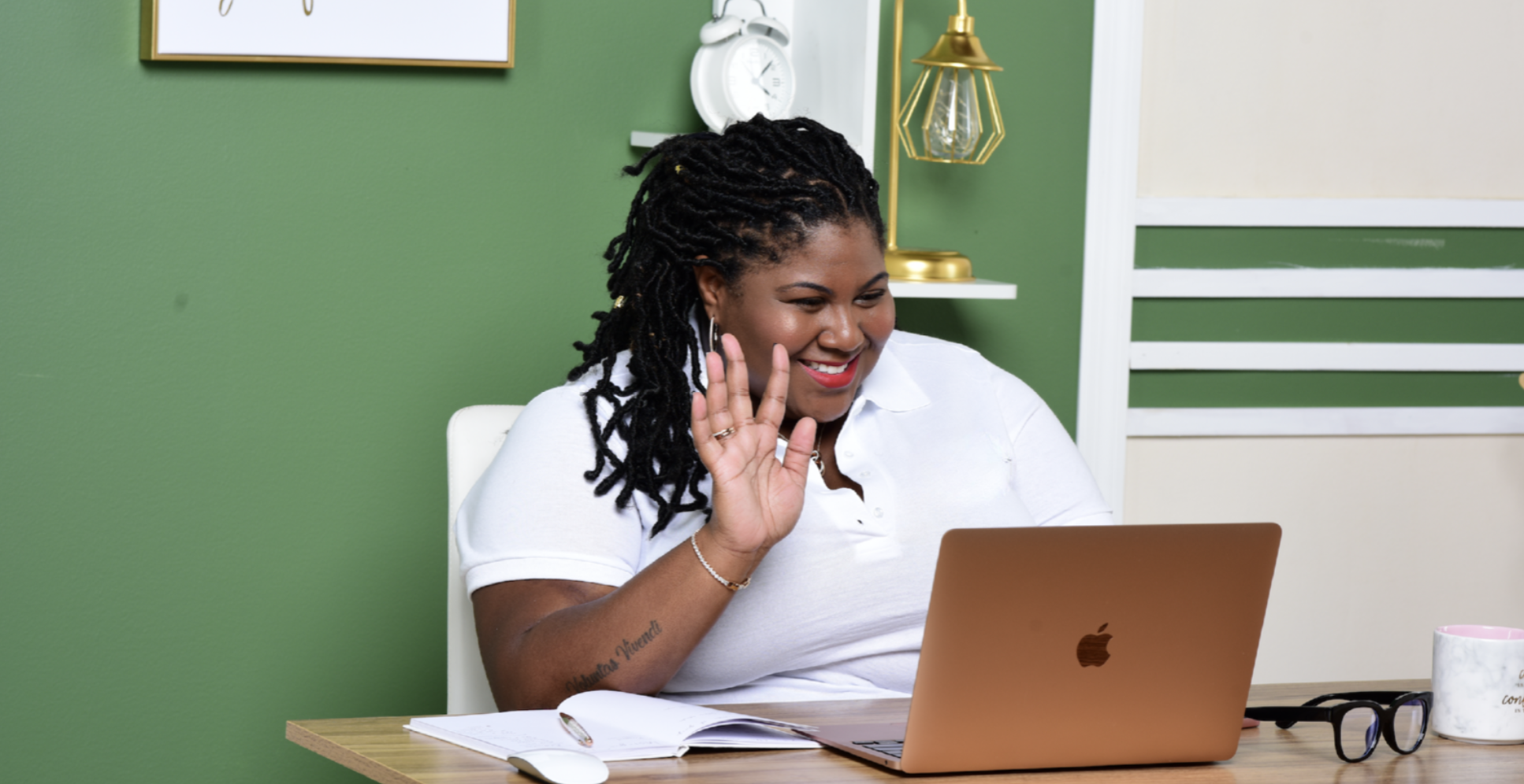 A woman with black locs smiles and waves at her laptop while seated at a desk. She wears a white polo shirt, earrings, and a bracelet.