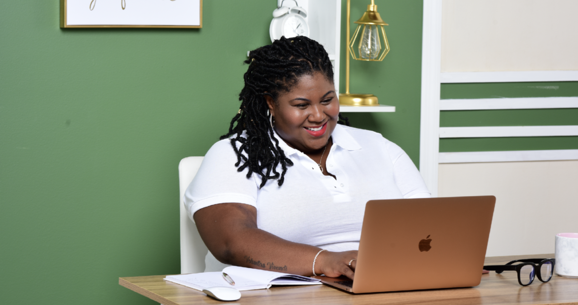 A woman with black, styled hair sitting at a desk, smiling while looking at a gold MacBook. She is wearing a white polo shirt, has a tattoo on her right arm, and is surrounded by a notebook, glasses, and a mug. The background features a green wall with a framed artwork, a white clock, and a gold lamp.