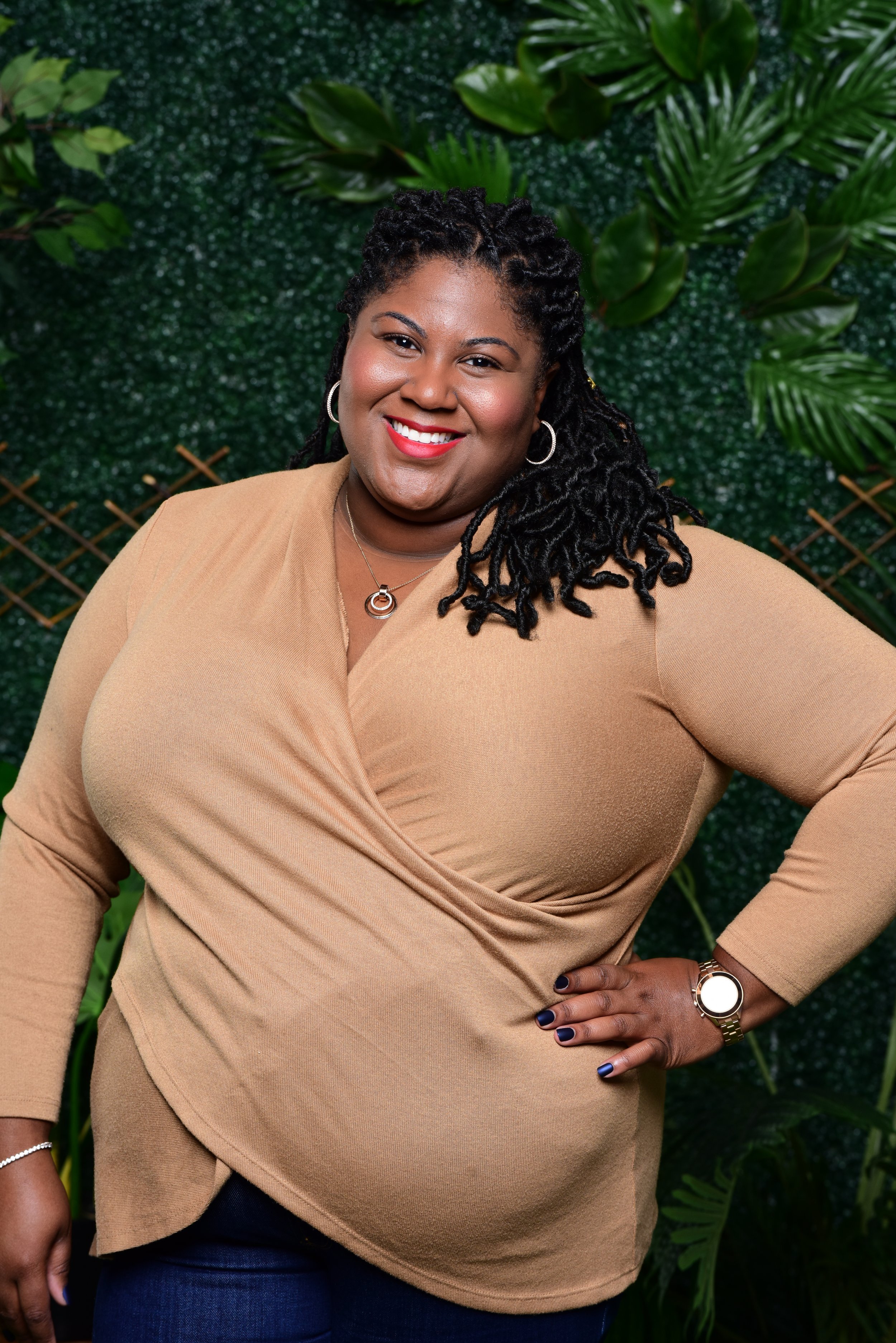 A woman smiling, wearing a beige top, dark pants, jewelry, and a watch, standing against a green leafy background.