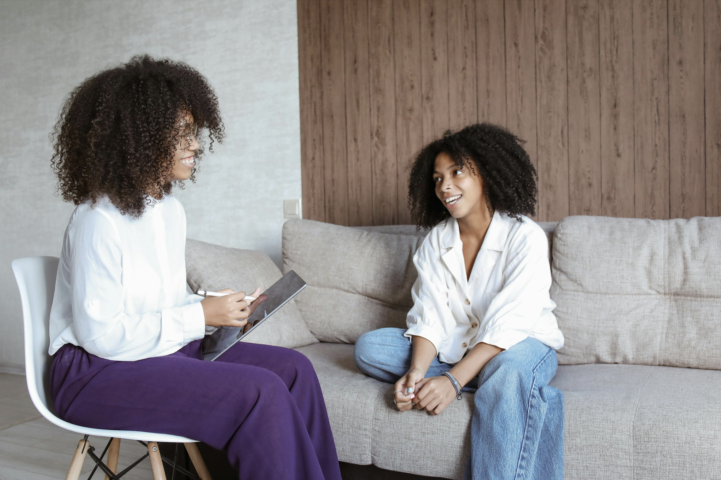 Two women with curly hair sitting and talking in a living room, one holding a tablet, smiling and engaging in conversation.