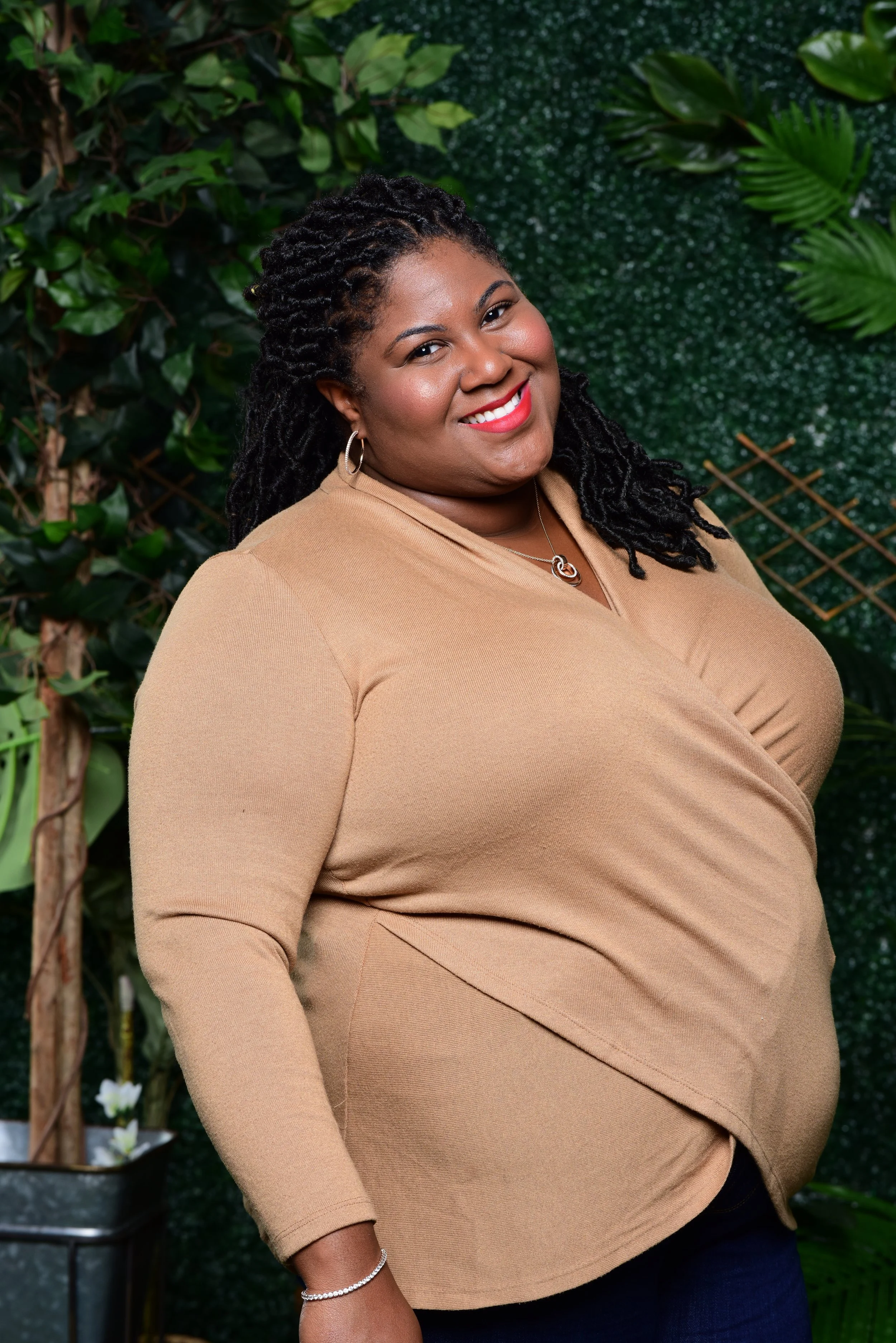 A woman with long locs, wearing a tan top and jewelry, standing in front of a green leafy backdrop, smiling at the camera.