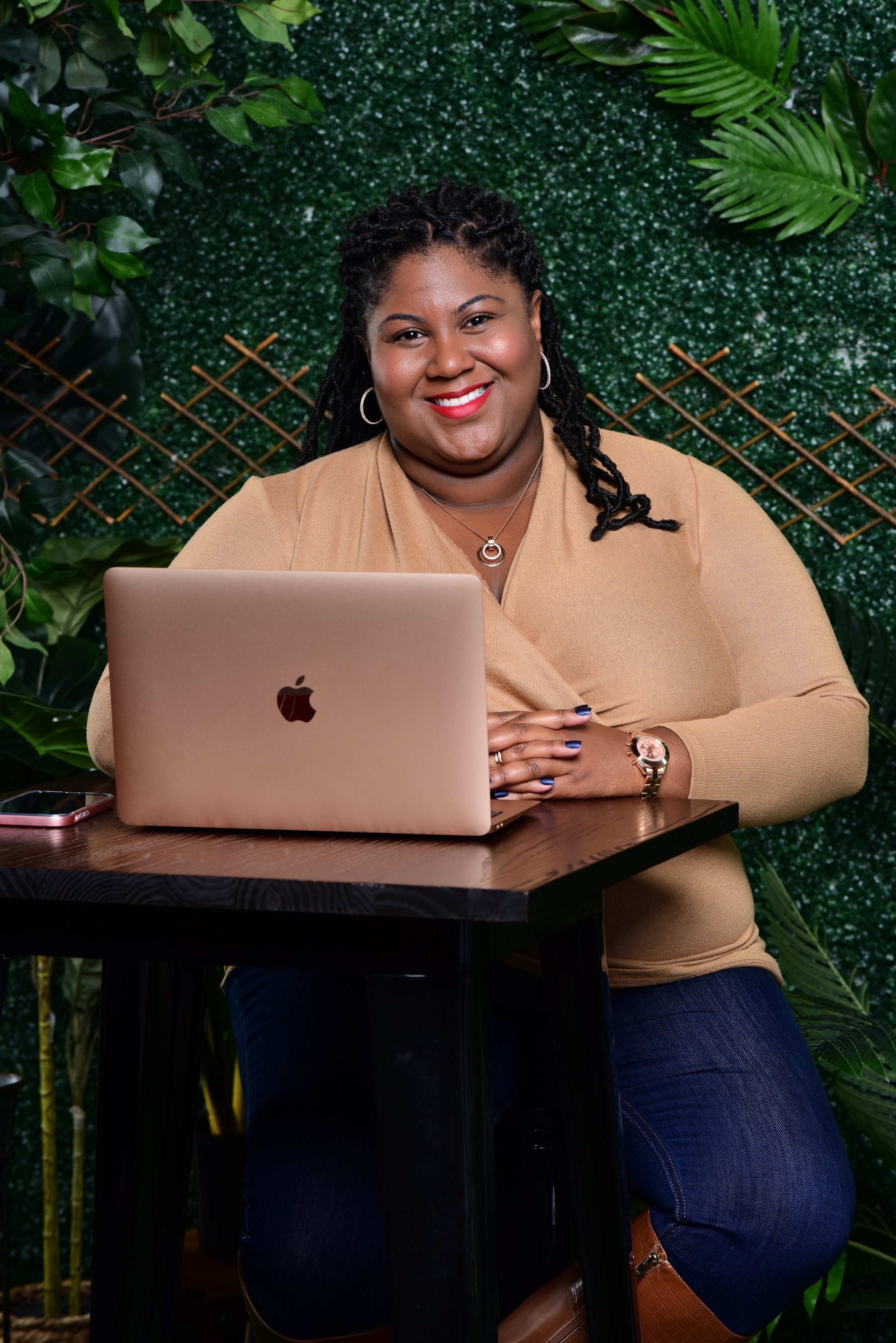 A woman with locs hair and red lipstick sitting at a wooden table with a silver Apple laptop, surrounded by lush green plants and a textured green wall background, smiling at the camera.