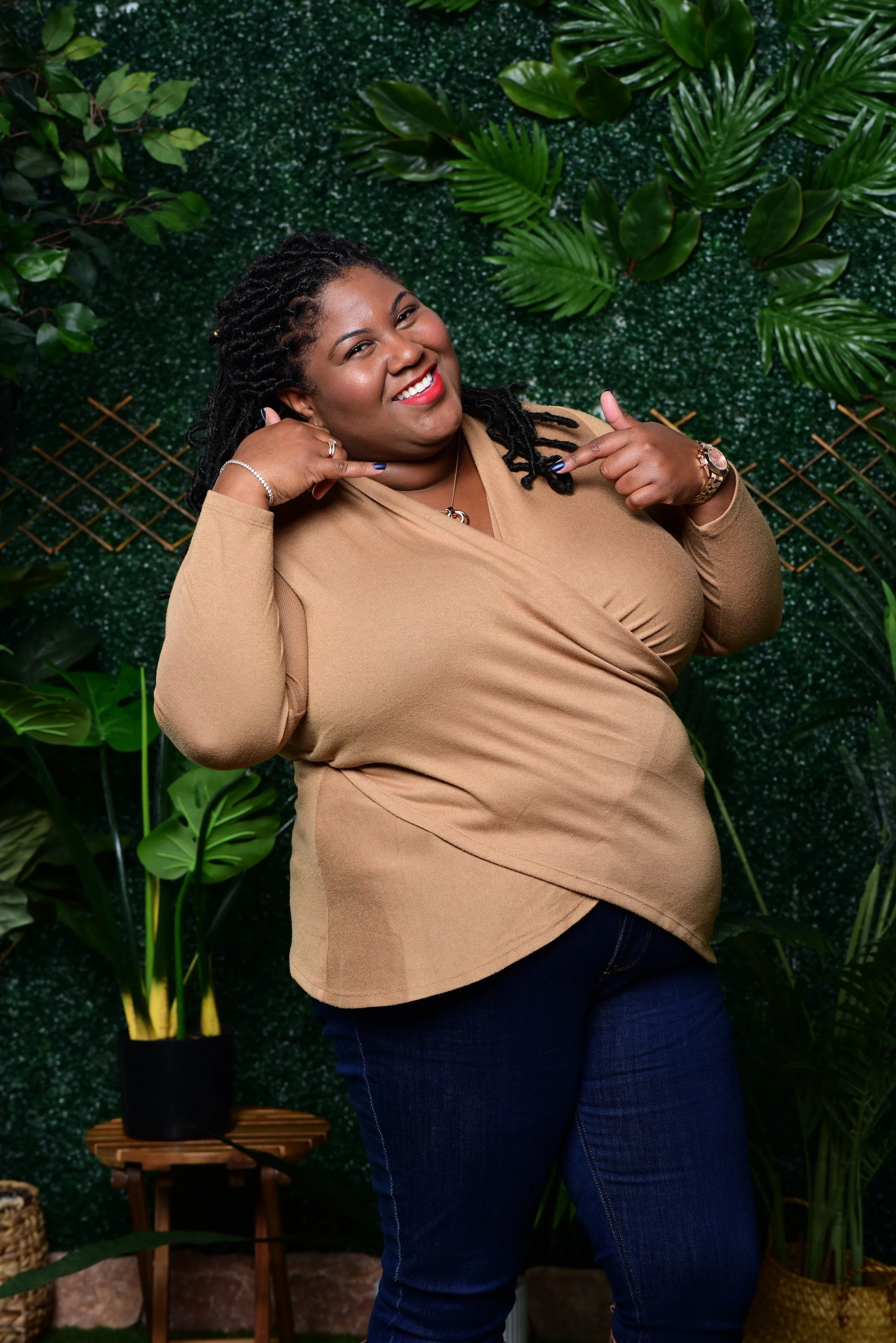 A smiling woman with dark curly hair wearing a tan long-sleeve top and dark jeans, posing confidently with her hands near her face, standing against a green botanical backdrop.