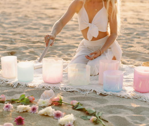 A woman in white clothing kneeling on a sandy beach, arranging pastel-colored candle holders on a cloth with flowers nearby at sunset.