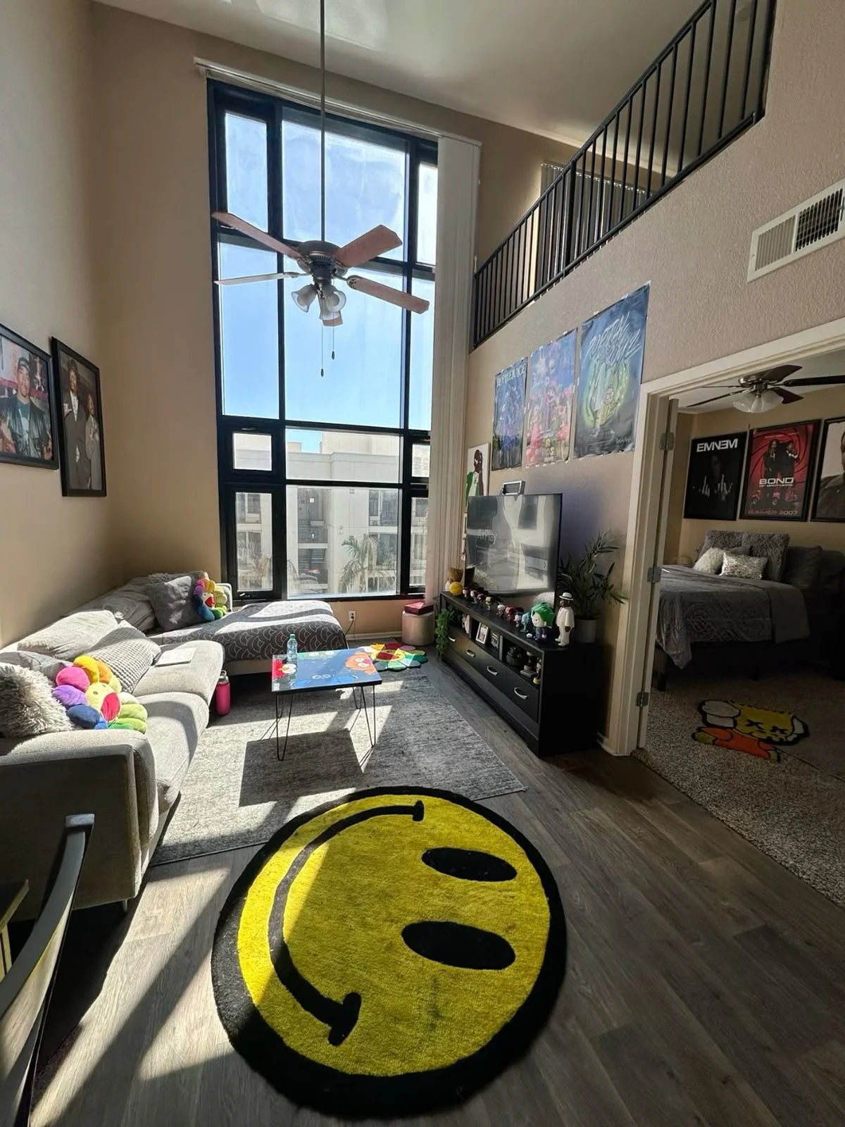 Living room with large window, ceiling fan, gray sofa with plush toys, coffee table, entertainment unit with decorations, colorful posters on the wall, and a doorway leading to a bedroom with framed posters on the wall. A smiley face rug is on the wooden floor.