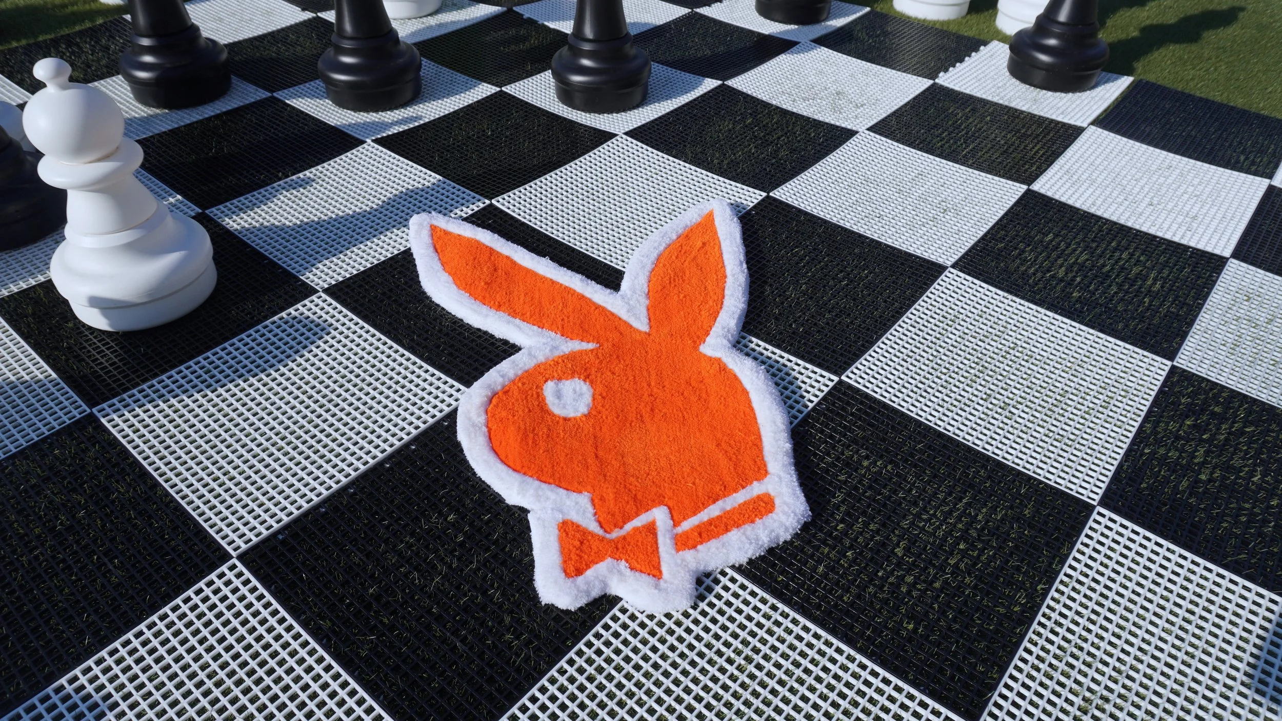 A chessboard set up outdoors with black and white pieces. A large orange and white bunny-shaped rug is in front of the board.