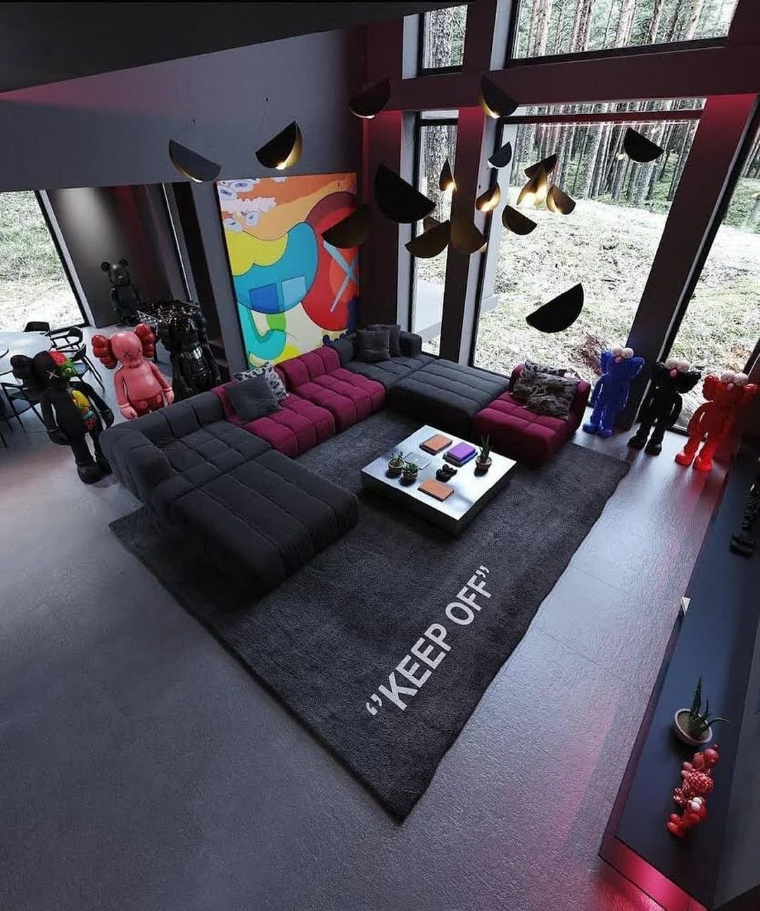 A modern living room with large floor-to-ceiling windows showing trees outside. The room has a dark gray sectional sofa with red cushions, a black-and-white rug with the words 'KEEP OFF,' a coffee table with books and plants, and colorful balloon sculptures of teddy bears and figurines. There is a large abstract painting on the wall and a contemporary black chandelier hanging from the ceiling.