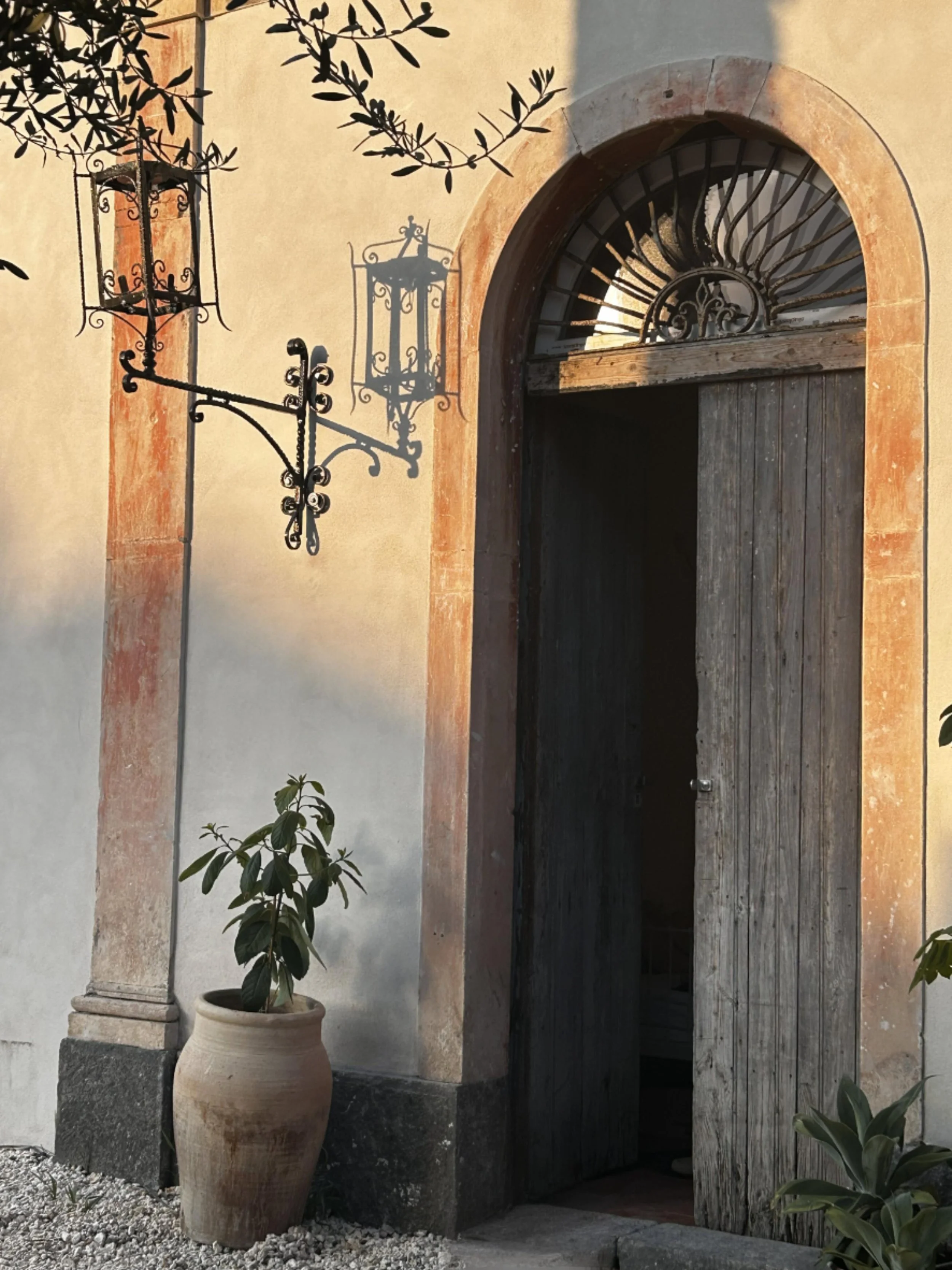 An old wooden door with a rounded archway at the top, framed by stone, partially open. There are two decorative wrought iron lanterns casting shadows on the wall to the left of the door. A large clay pot with a potted plant is on ground level in fron