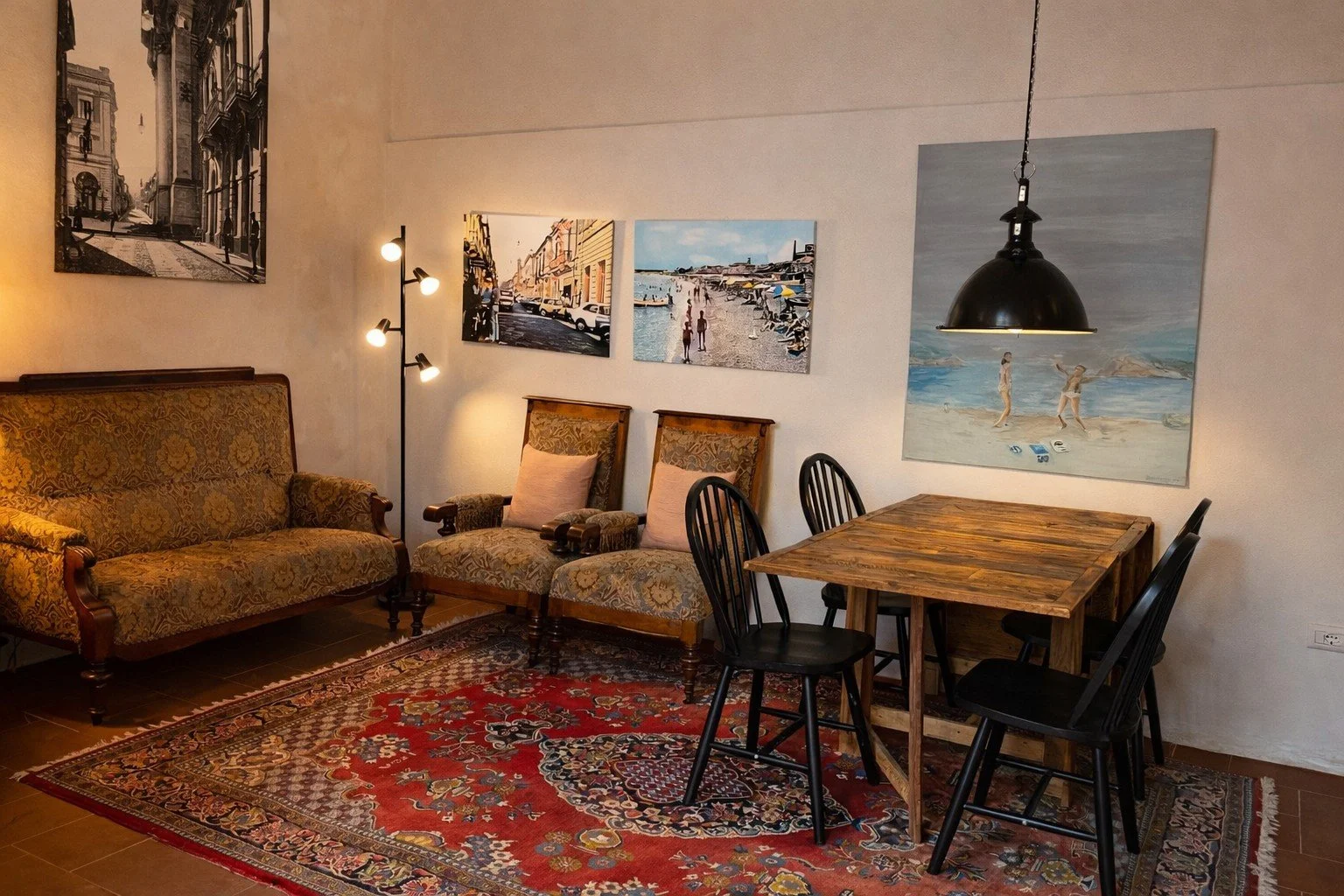 Living room with antique sofa and two chairs, a wooden dining table with four black chairs, colorful wall art, a red patterned rug, and a black pendant light.