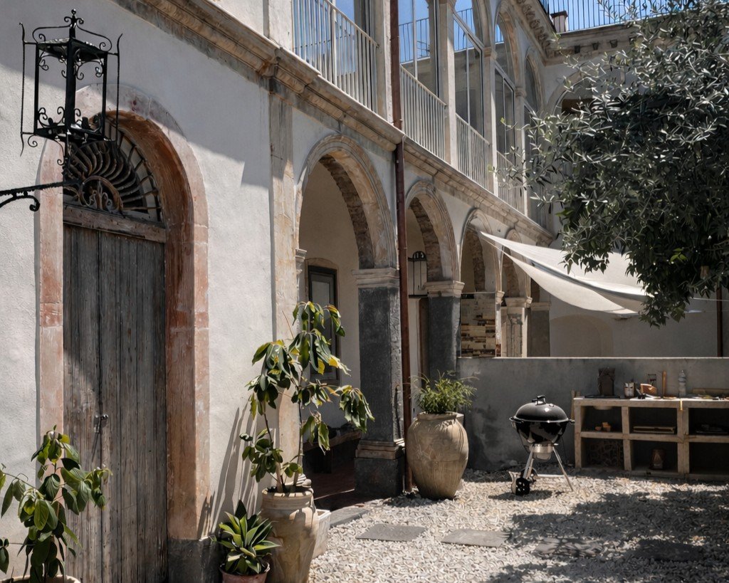 A courtyard with potted plants, a barbecue grill, and a rustic stone building with arches and balcony railings.