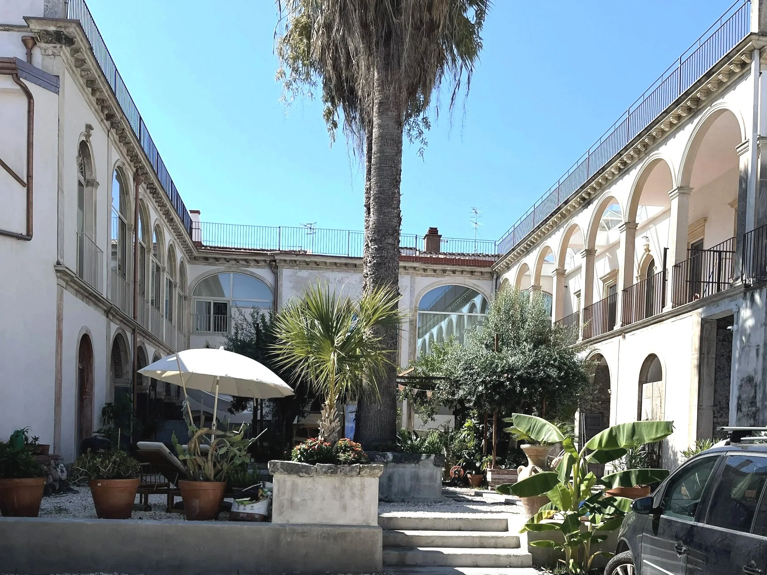 Courtyard with potted plants, a large palm tree, an umbrella, and a parked car, surrounded by white buildings with arched windows and railings on the balconies under a clear blue sky.