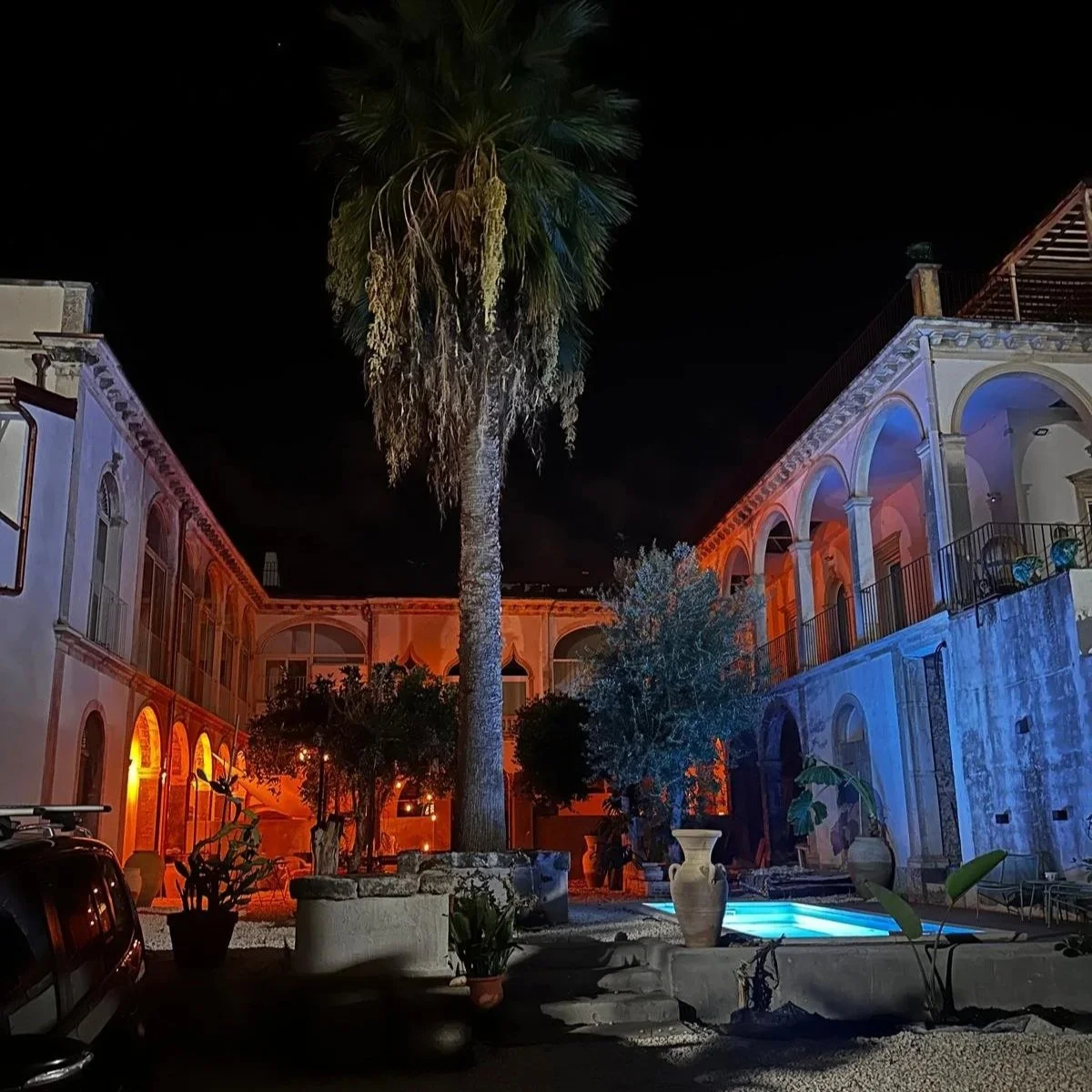 A nighttime scene of a courtyard with colorful lighting. There is a tall palm tree in the center, surrounded by potted plants and a small swimming pool. The courtyard is enclosed by a multi-story building with arched windows and balconies, illuminate