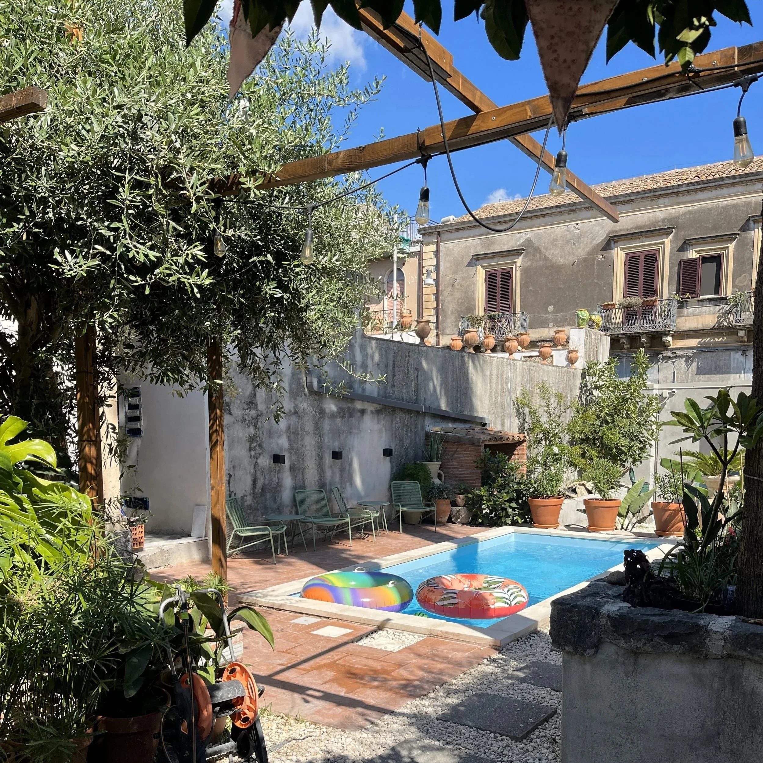 A private outdoor patio with a swimming pool, surrounded by potted plants and trees. There are two colorful pool floats in the pool, metal chairs along the wall, and string lights overhead. An old building with windows and balconies is visible in the