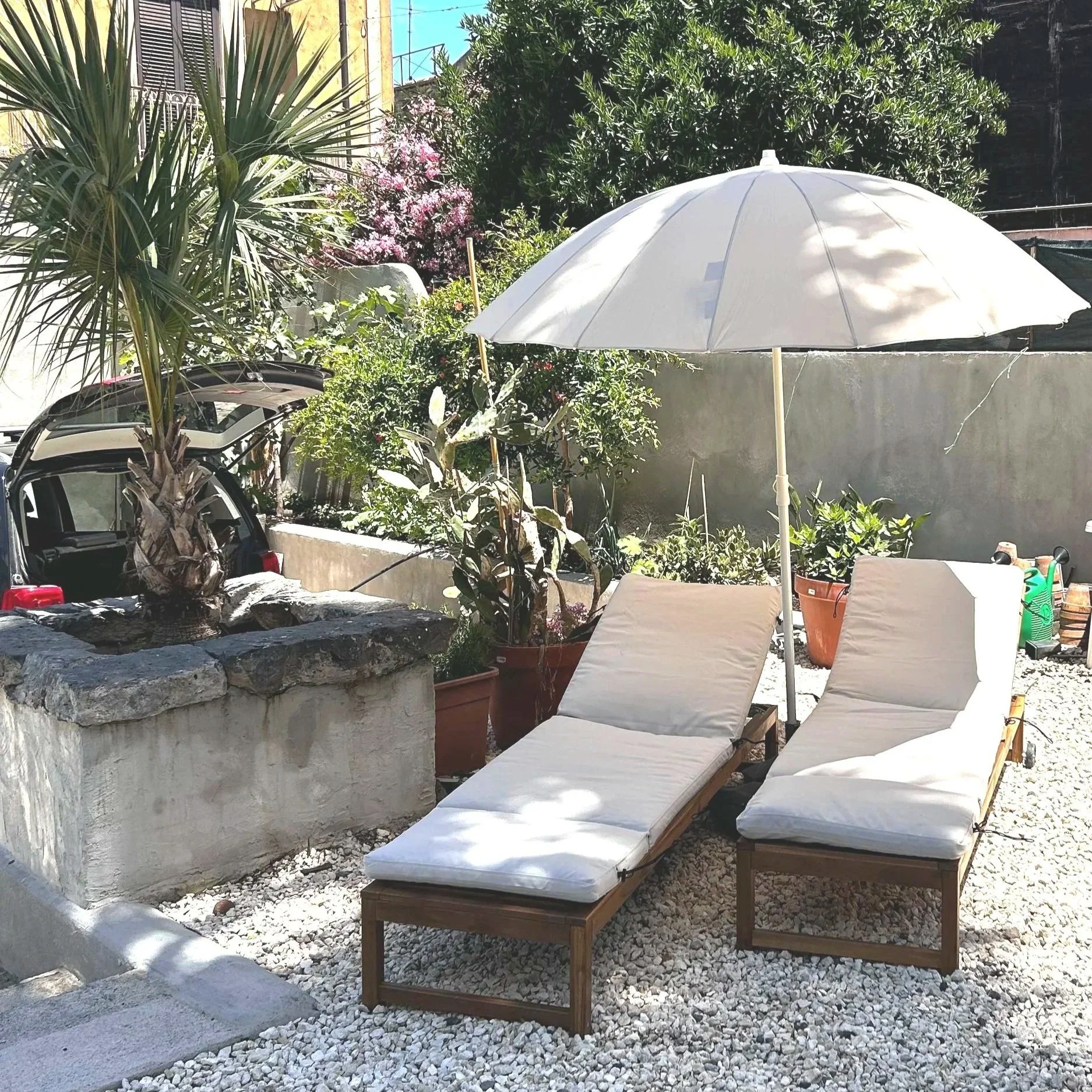 Two white cushioned lounge chairs under a large white patio umbrella in a garden with potted plants, trees, and a stone planter. A car with an open trunk is visible in the background.