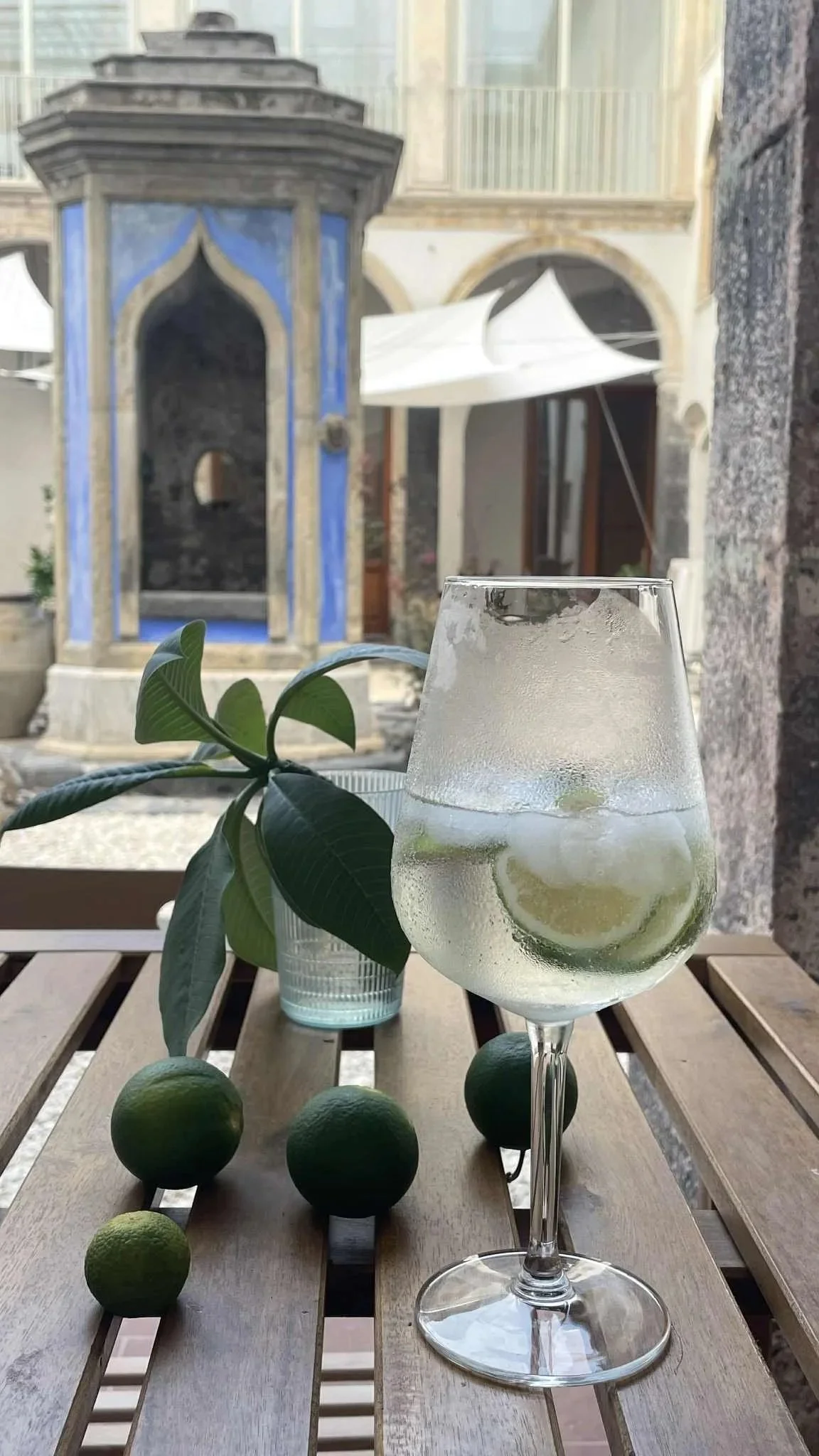 A glass of fizzy lemonade with ice and lemon slices on a wooden table, with limes and a potted plant nearby, in an outdoor patio with a stone fountain and Mediterranean-style architecture in the background.