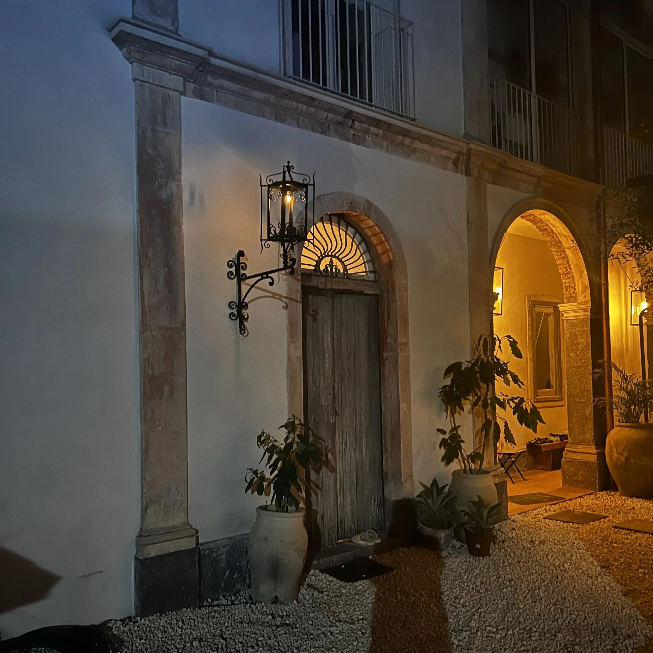 Nighttime exterior of a house with a rustic wooden door, illuminated by warm yellow lights. Potted plants and decorative elements are arranged near the entrance, with a wrought iron lantern hanging on the wall.