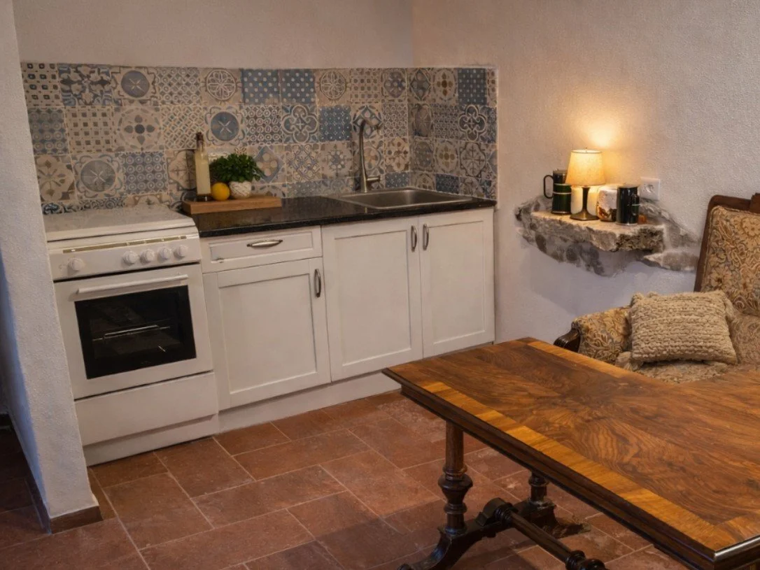 Small kitchen with white cabinets, a black countertop, a colorful patterned tile backsplash, a small sink, and a stove. There is a wooden cutting board with greenery and lemons, and a shelf with a lamp and mugs. A wooden table and an upholstered armchair with a decorative pillow are also visible.