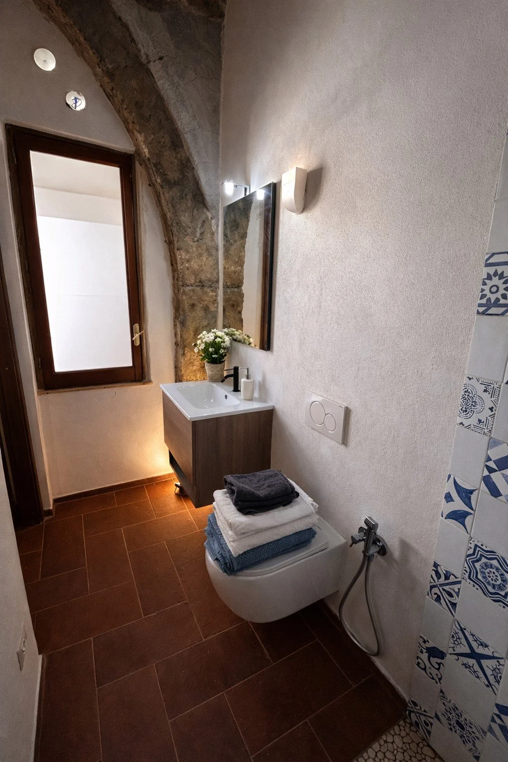 Small bathroom with a wooden vanity, white sink, flowers, mirror, window, and folded towels on the toilet. Exposed stone wall and patterned tiles on the right.