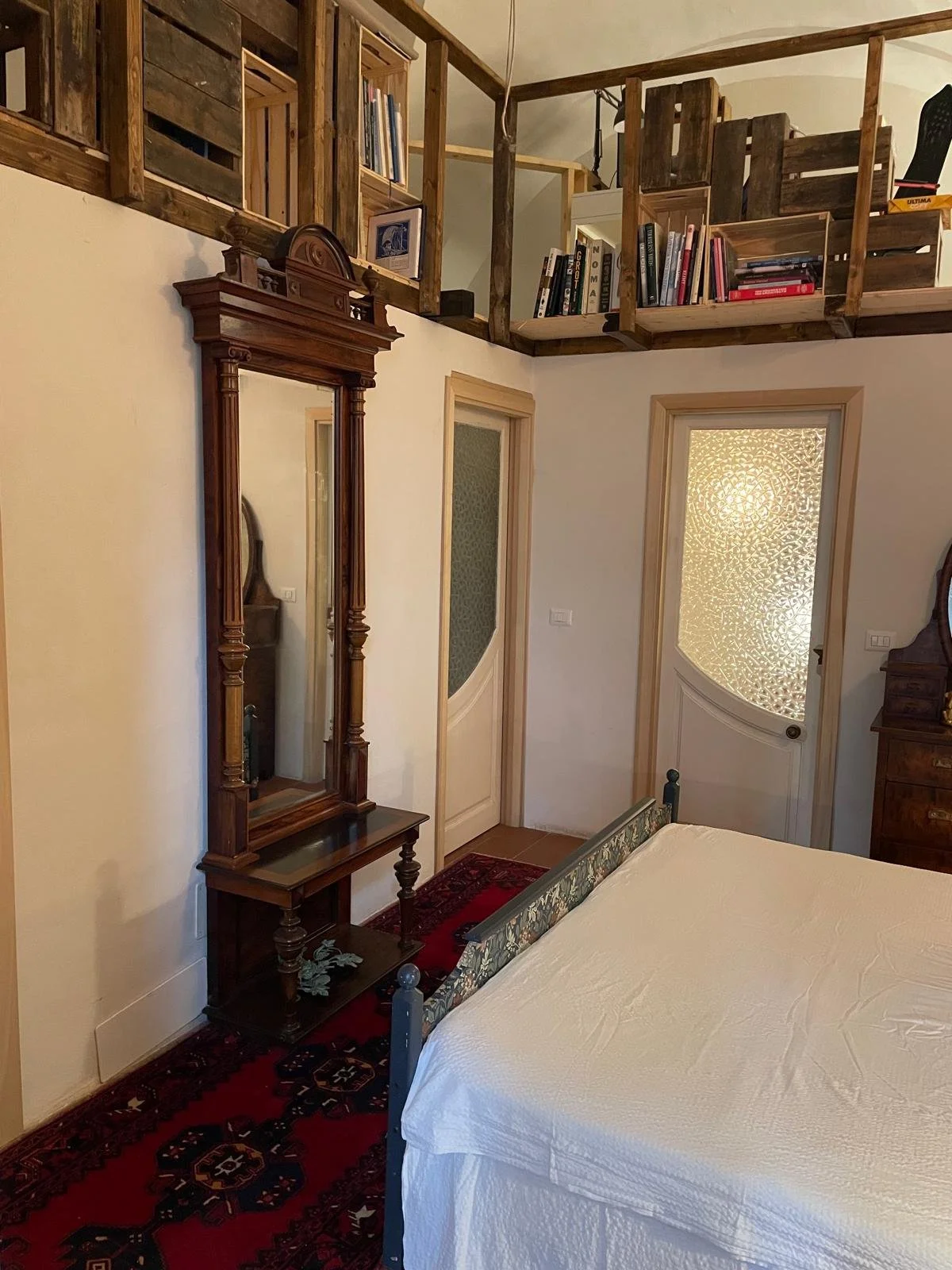 A bedroom with antique furniture and a loft area with books. A wooden mirror with a small table, a bed with a floral headboard, a cabinet, and two doors with textured glass panels. The loft is built with wooden pallets and contains books.