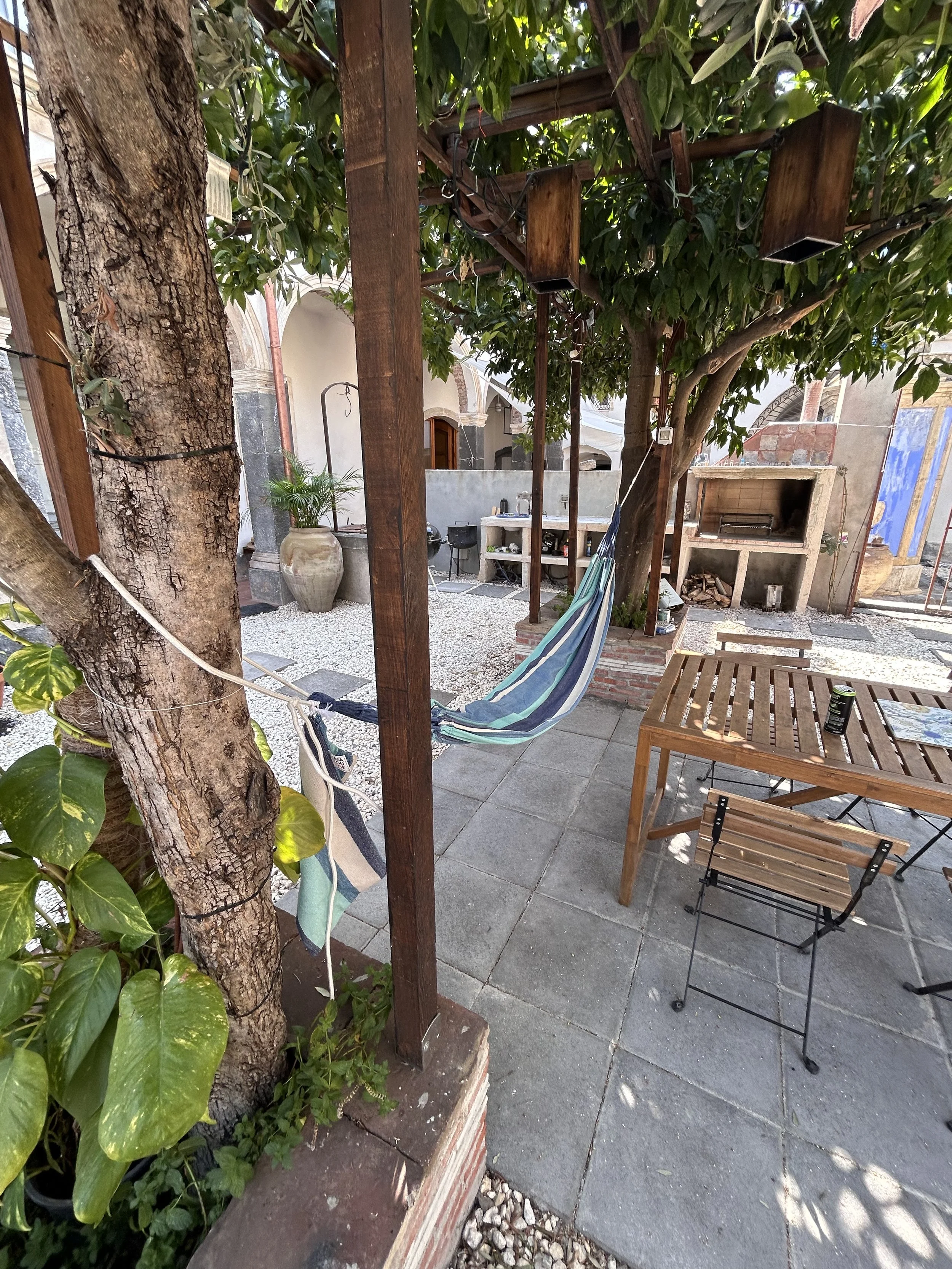 An outdoor patio area with a hammock tied between a tree and a wooden post, surrounded by potted plants, a wooden table with a folding chair, and a grill in the background.