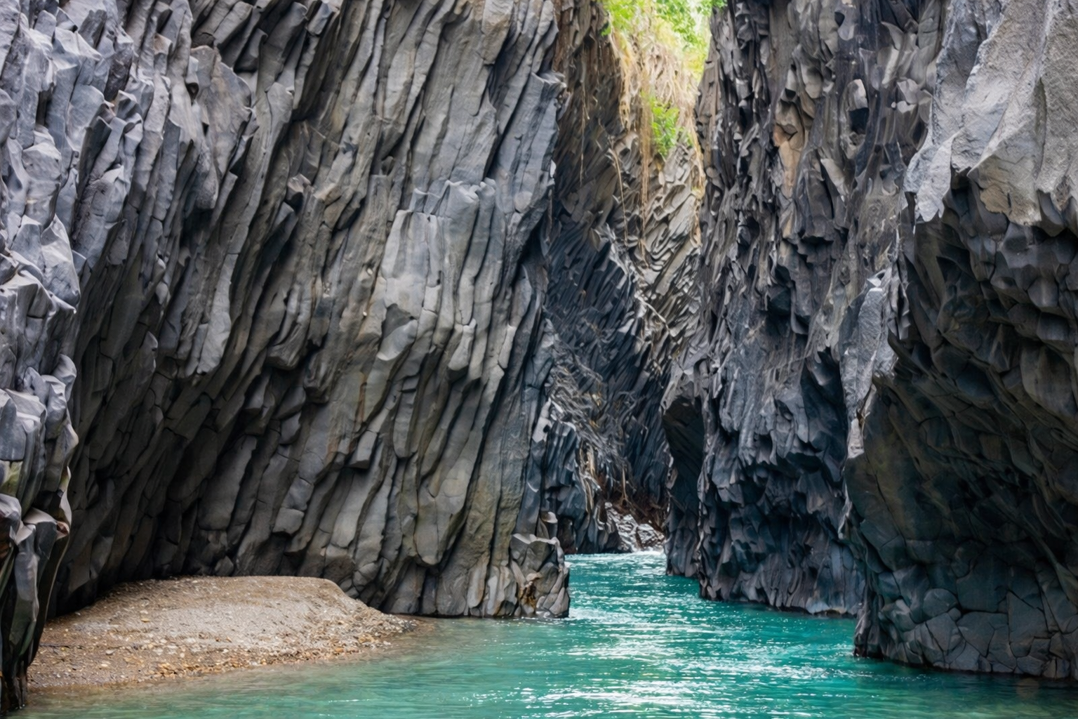 A narrow canyon with steep, jagged walls made of layered gray rocks flanking turquoise water at the bottom.