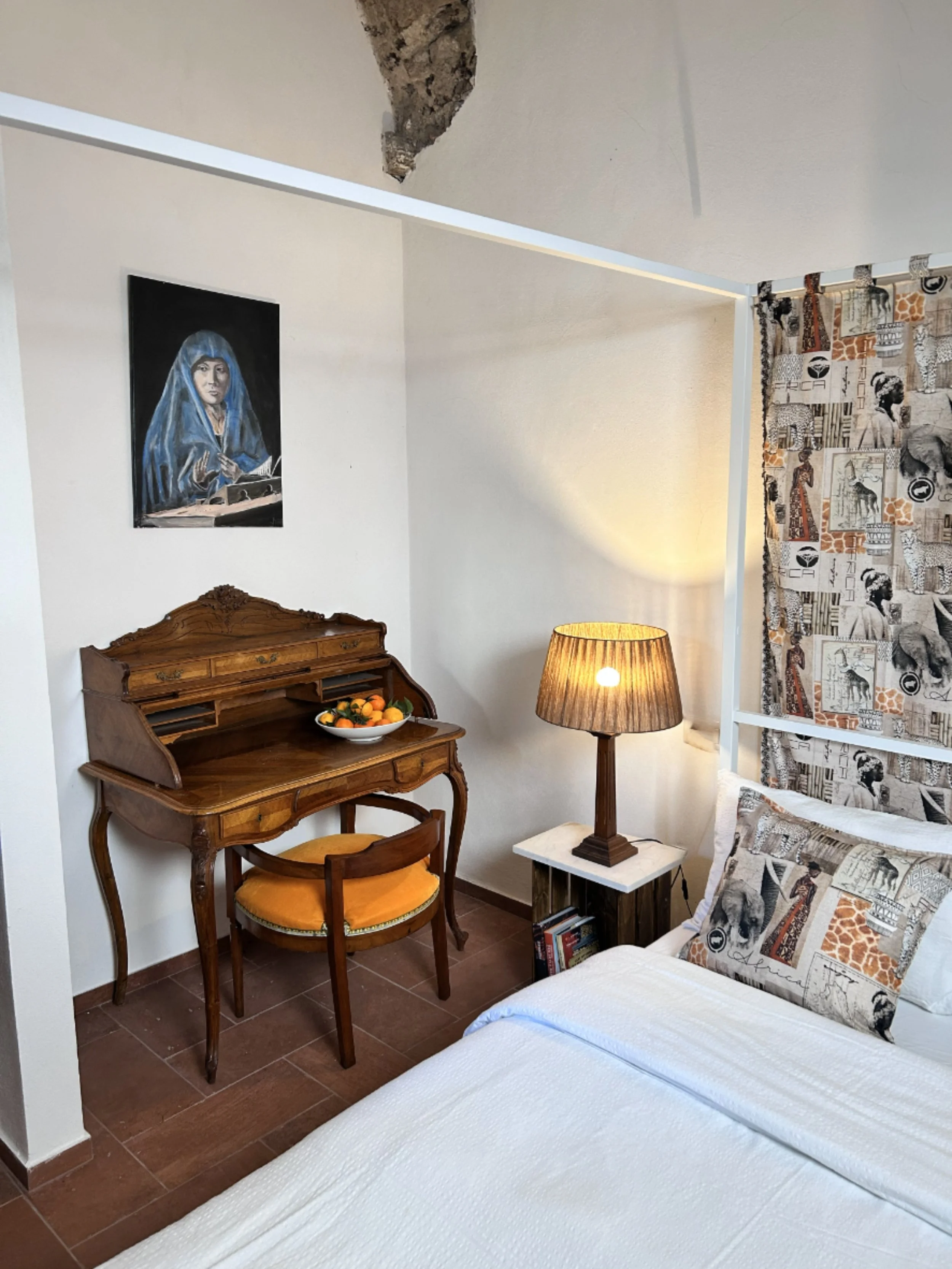 A cozy bedroom corner with a bed, decorative pillows, a bedside table with a lamp, a vintage wooden writing desk with a bowl of fruit, and wall art depicting a woman.