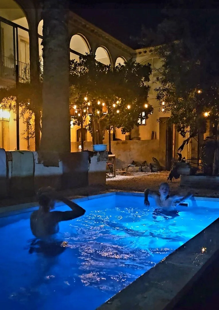 Two children are swimming and playing in a brightly lit swimming pool at night, surrounded by trees and illuminated string lights, near a building with arched windows.