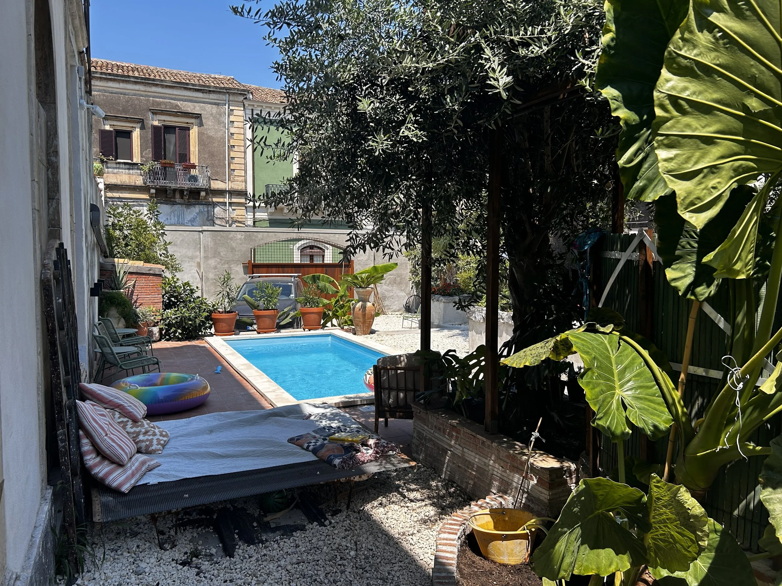 A backyard with a swimming pool surrounded by potted plants, outdoor furniture, and shaded by trees, with residential buildings in the background.