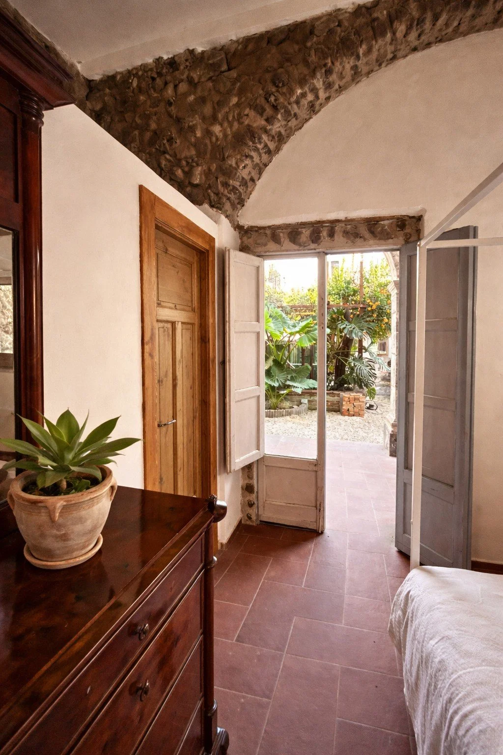 A cozy room with a rustic stone arch ceiling, a wooden door, and open white shutters revealing a lush garden outside. There is a dark wooden dresser with a potted succulent plant, and part of a bed with a white blanket is visible.