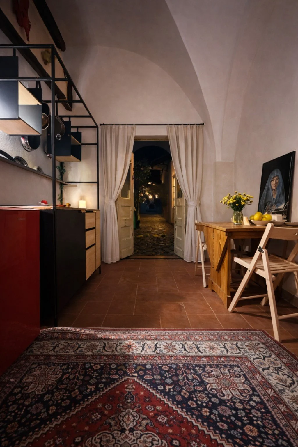 Interior of a cozy room with a view outside through an open door, featuring a patterned rug, a wooden table with a vase of flowers, a chair, framed artwork, curtains, built-in shelves with dishes, and warm wall lighting.