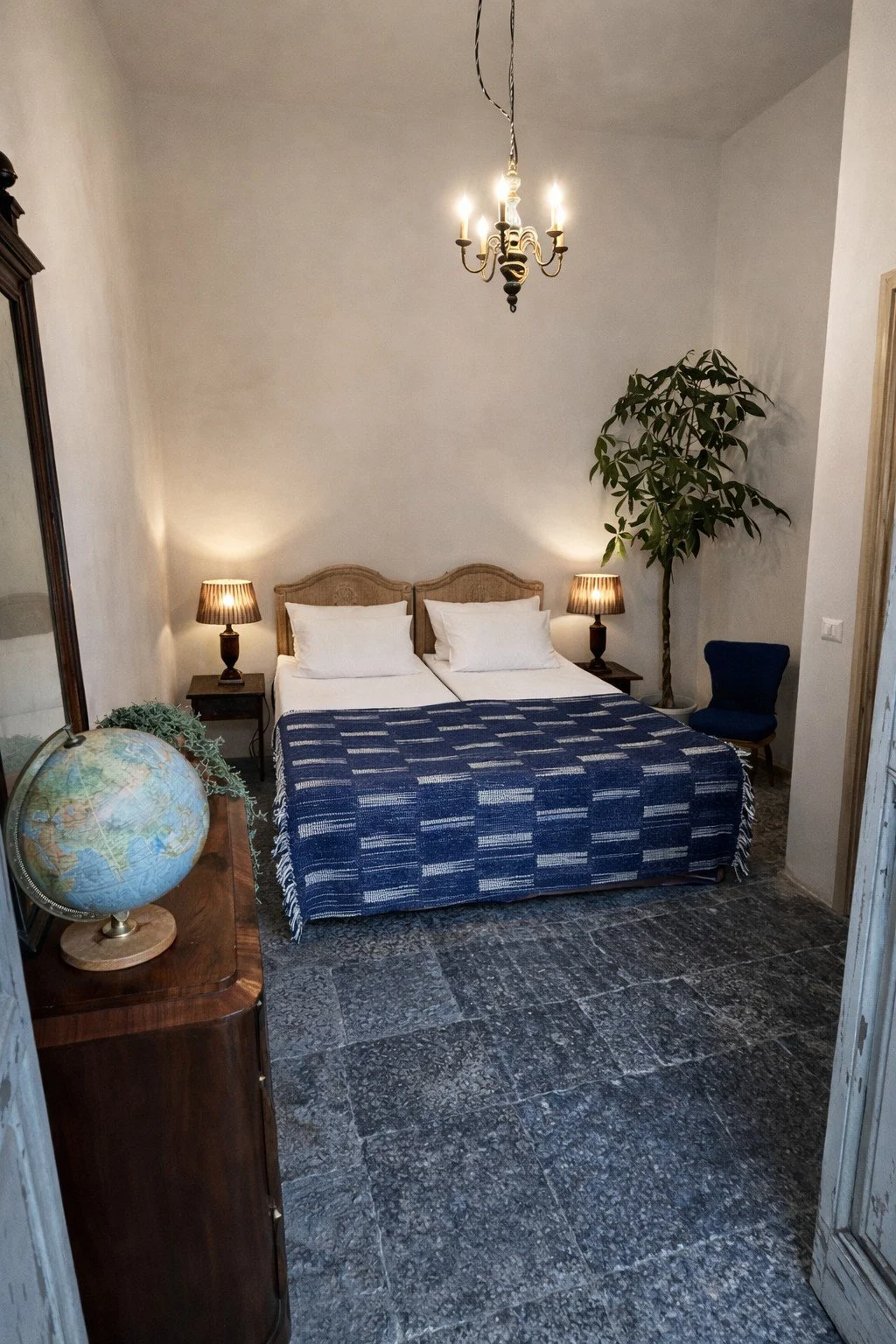 A cozy bedroom with a double bed, white pillows, and a blue striped blanket. Two small nightstands with lamps flank the bed. A globe sits on a wooden cabinet to the left, and a blue armchair is in the corner near a tall plant. A chandelier hangs from the ceiling, illuminating the room.