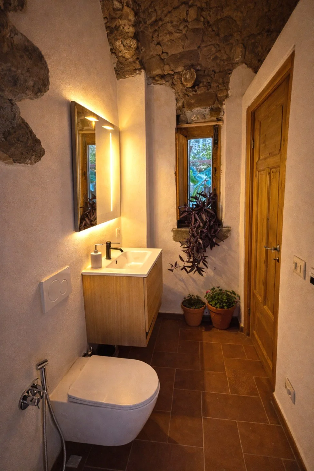 A small bathroom with rustic stone and wood accents, including a white wall-mounted toilet, a wooden cabinet with a sink, a mirror with backlit lighting, a narrow window with plants outside, and two potted plants on the floor.