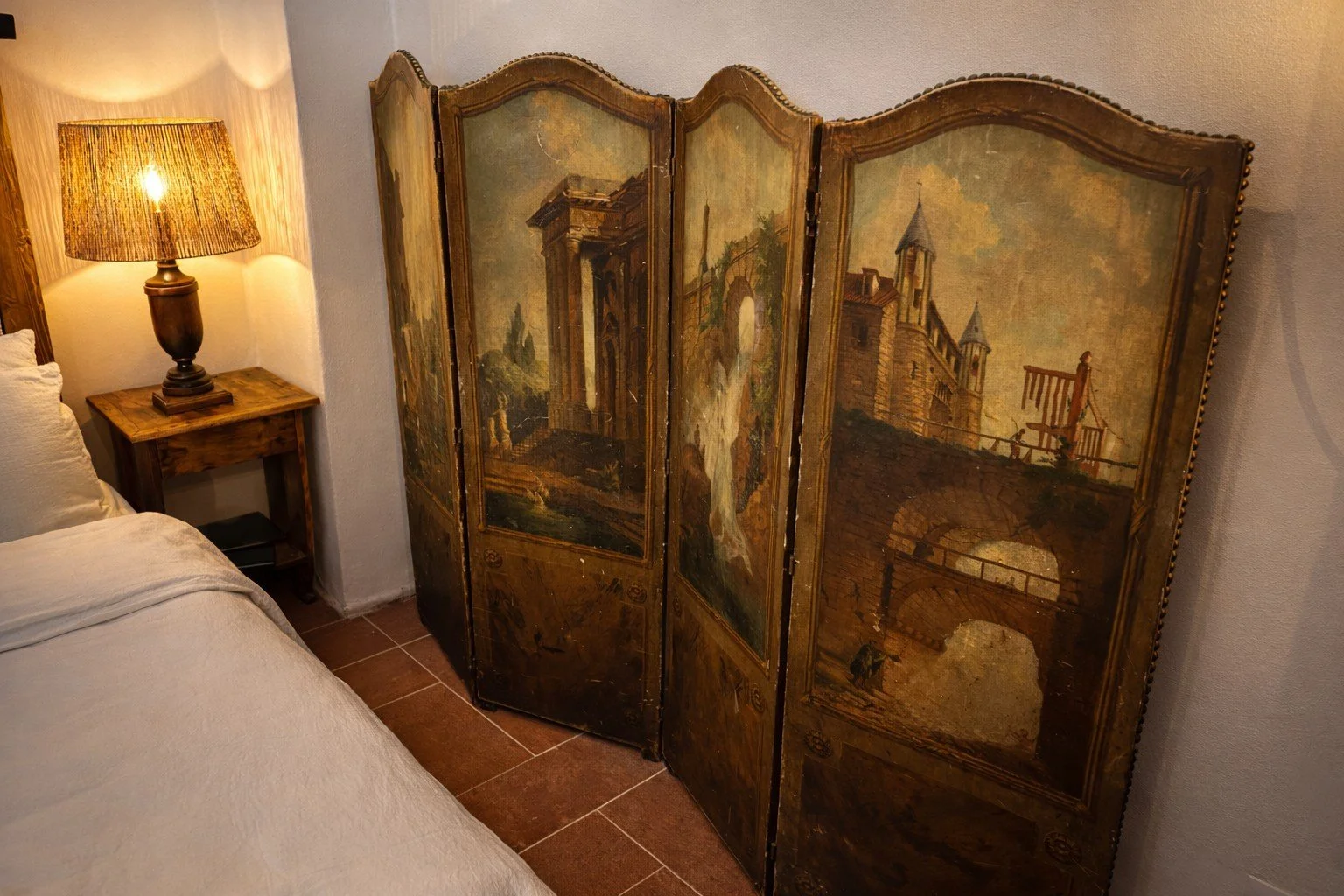 A vintage painted folding room divider featuring landscape scenes with classical architecture and bridges, positioned beside a bed with white bedding and a wooden nightstand with a lamp, in a cozy bedroom.