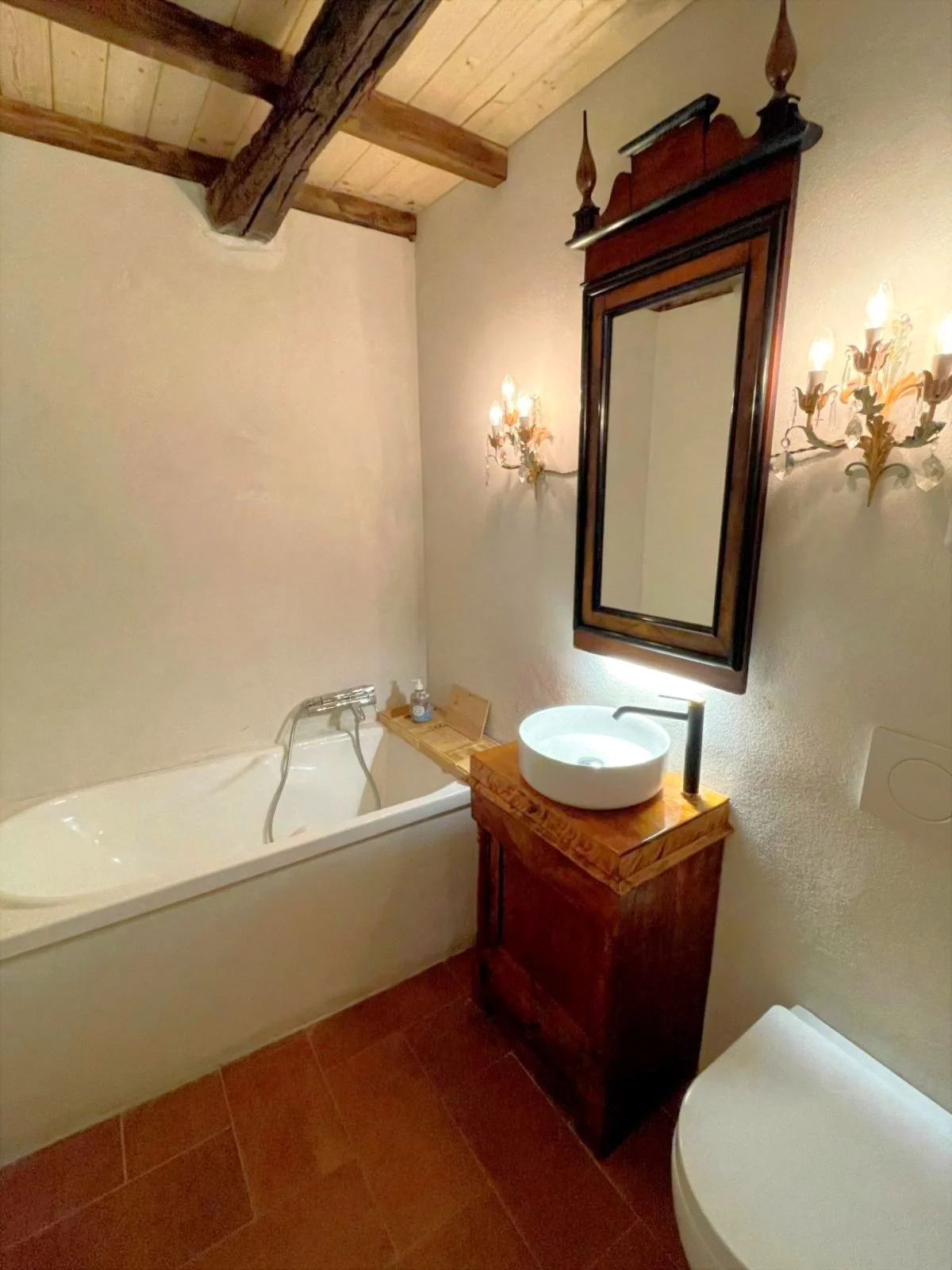 Bathroom with a bathtub, a wooden vanity with a round vessel sink, a mirror, and ornate wall sconces, with a wooden ceiling and tiled floor.