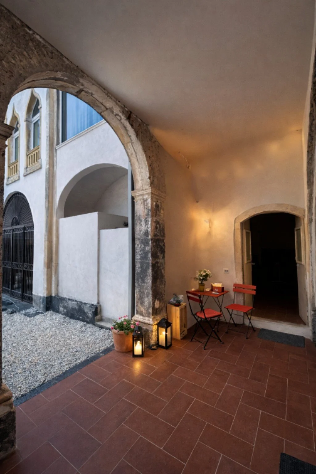 Courtyard with terracotta tile floor, potted flowers, two lanterns, and a small table with two chairs near an arched doorway, with soft lighting on the wall.