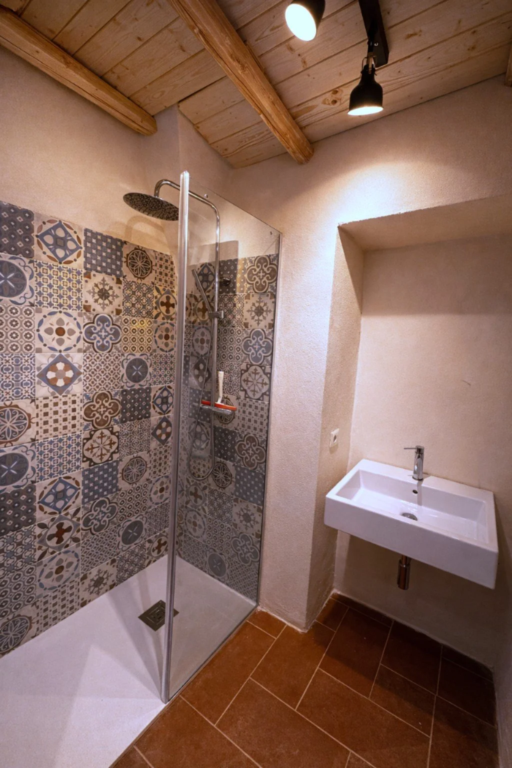 A small bathroom with a corner shower featuring patterned tiles, a white wall-mounted sink, and a wooden ceiling with track lighting.