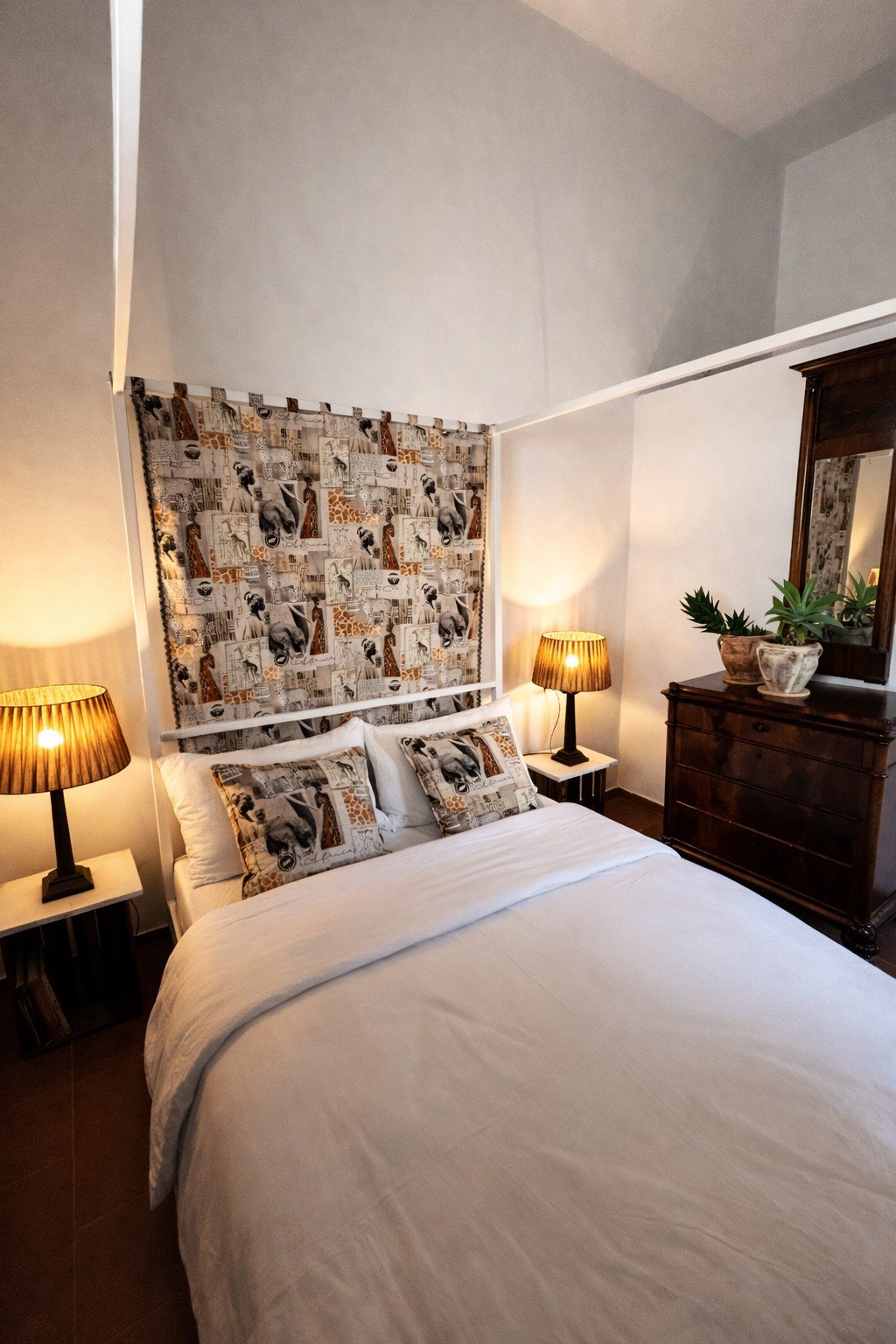 A cozy bedroom with a white bed, animal print pillows, and a matching animal print fabric on the wall behind the bed. There are two bedside tables with warm-lit table lamps, a wooden dresser with potted plants, and a mirror.