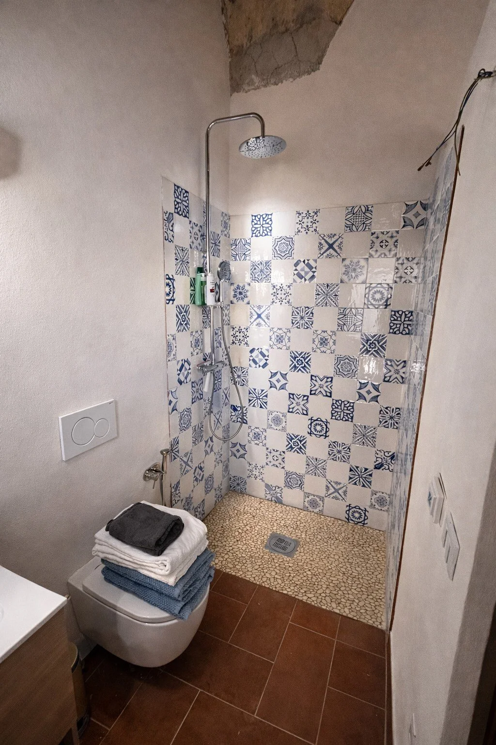 A small bathroom with a walk-in shower with patterned blue and beige tiles, a rain showerhead, a handheld shower, and shelves holding toiletries, next to a wall-mounted toilet with a stack of folded towels on top, and a brown tile floor.
