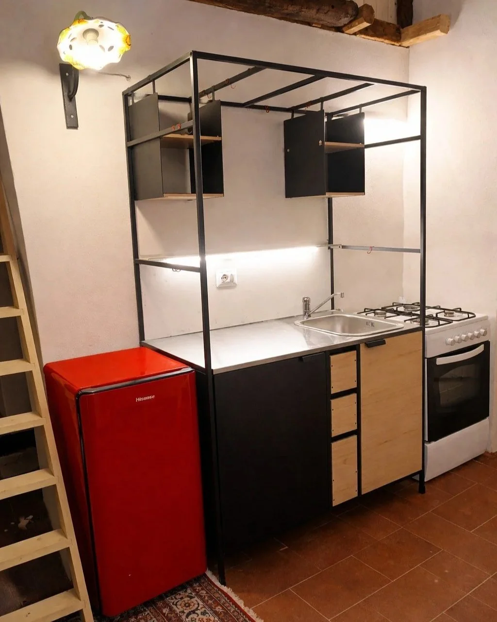 Compact kitchen setup with black metal frame, open shelves, beige and black cabinets, a stainless steel sink, a white stove, and a red mini refrigerator. Walls are white, and there is a decorative ceiling light.
