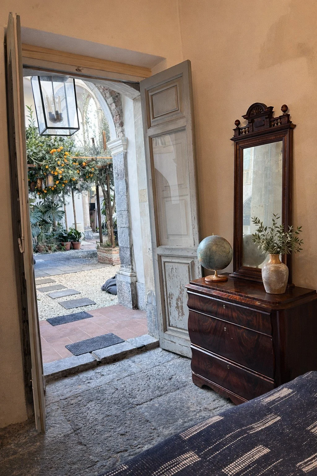 Living room with a wooden dresser, mirror, globe, and vase with greenery, open door leading to outdoor patio with orange trees and potted plants.