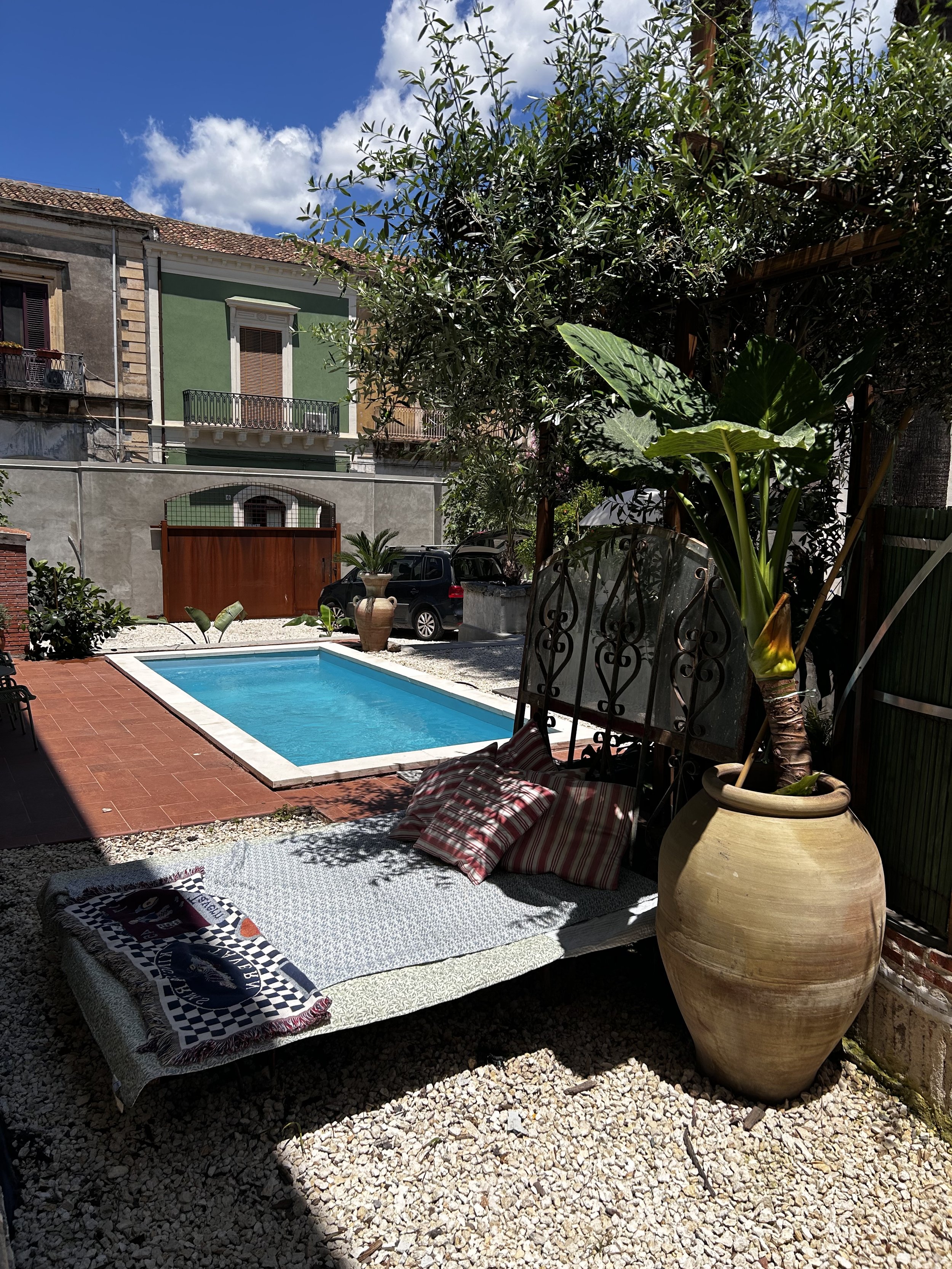 A backyard with a swimming pool, a sunbed with pillows and a towel, large potted plants, trees, and surrounding buildings under a partly cloudy sky.