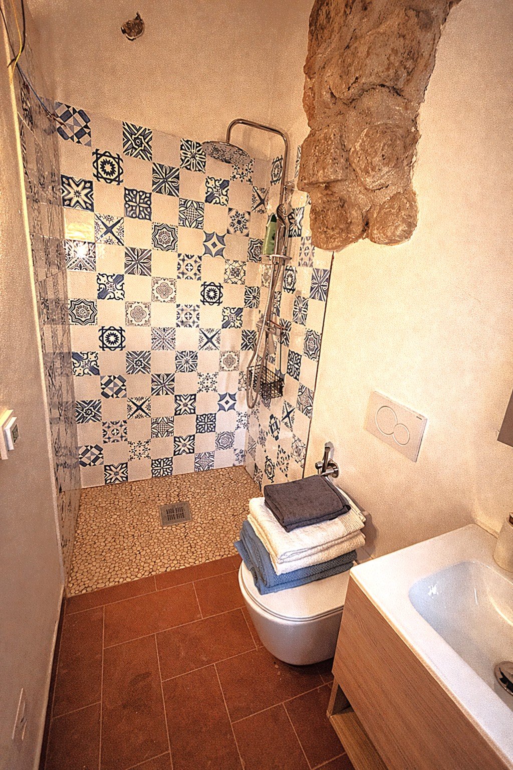 Bathroom with a shower area adorned with patterned blue and white tiles, a small pebble-tiled floor in the shower, a stack of folded towels on a toilet, and a wooden vanity with a sink. There is a stone wall feature and a flush plate on the wall.