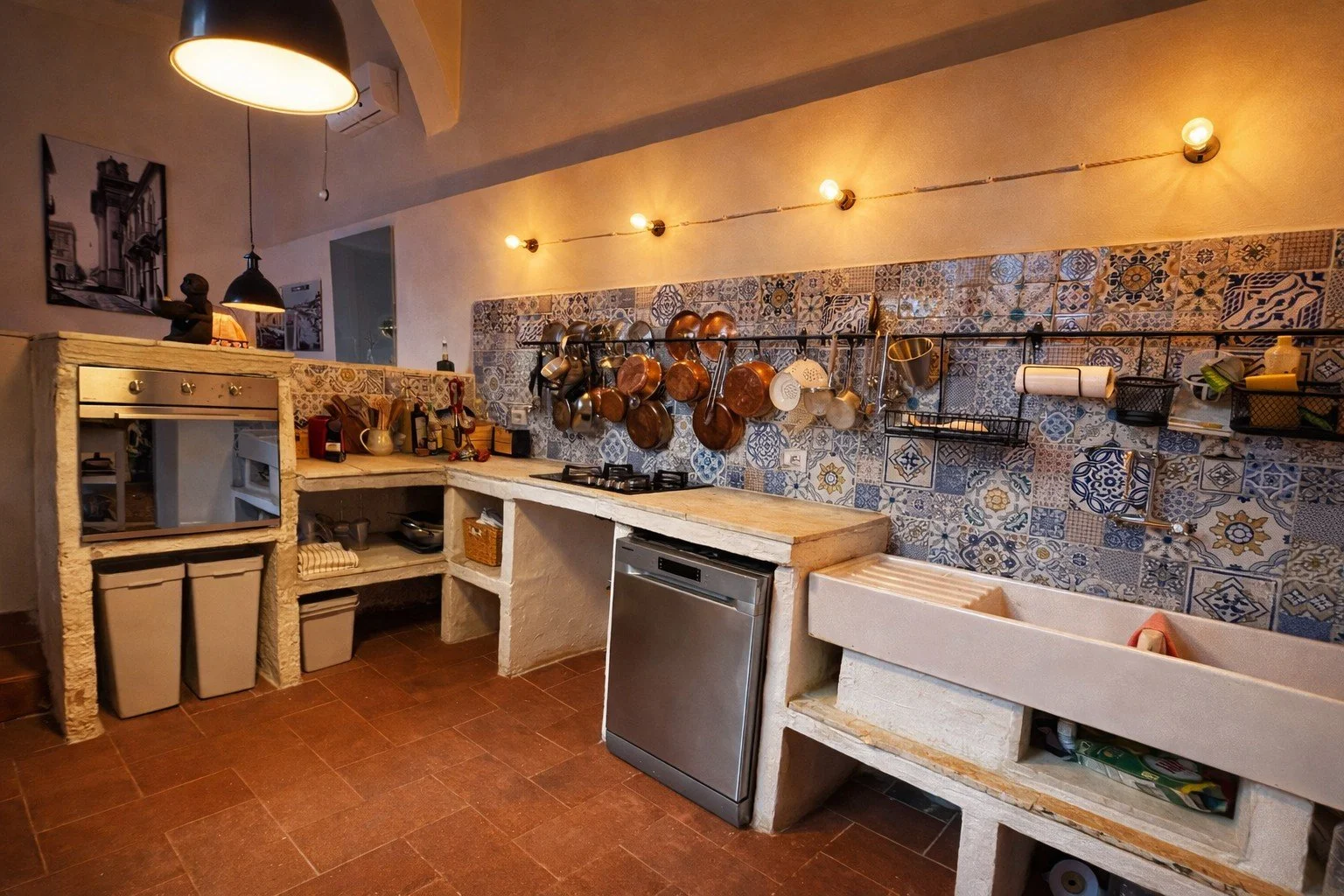 A rustic kitchen with a white stone counter, copper and stainless steel pots hanging on a rod, a small dishwasher, and a white utility sink, decorated with colorful patchwork tile backsplash, warm lighting, and wall art.