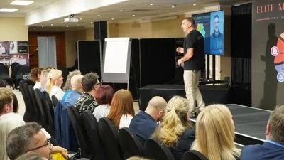 A speaker presenting to an audience in a conference room, with a large screen displaying a headshot and presentation materials.