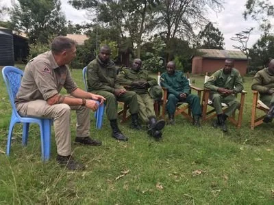 Six men are sitting outdoors on chairs and benches, engaged in a conversation. One man, dressed in a tan uniform, sits on a blue plastic chair, while the other five men, dressed in military uniforms, are seated on wooden benches. The setting is grassy with trees and buildings in the background.