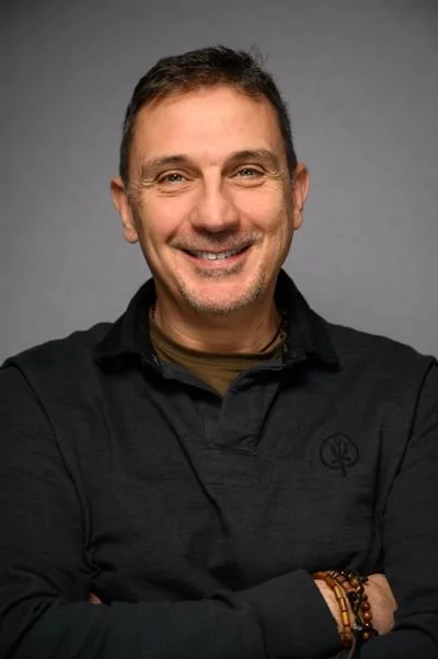 Portrait of a smiling man with short dark hair, light beard, wearing a black jacket with a logo, and a bracelet on his wrist, standing against a gray background.