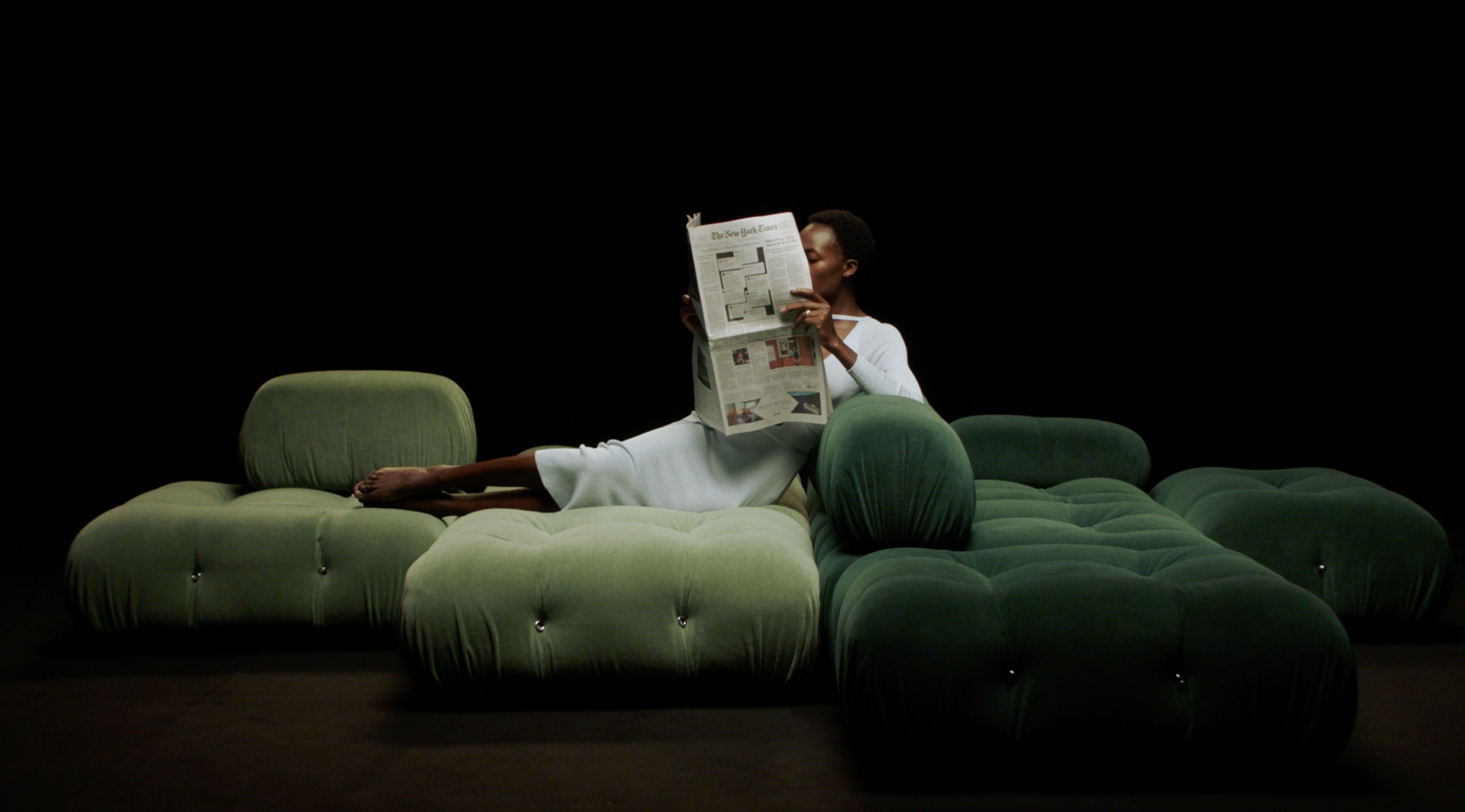 Donna seduta su un divano verde, che legge il giornale in un ambiente scuro.