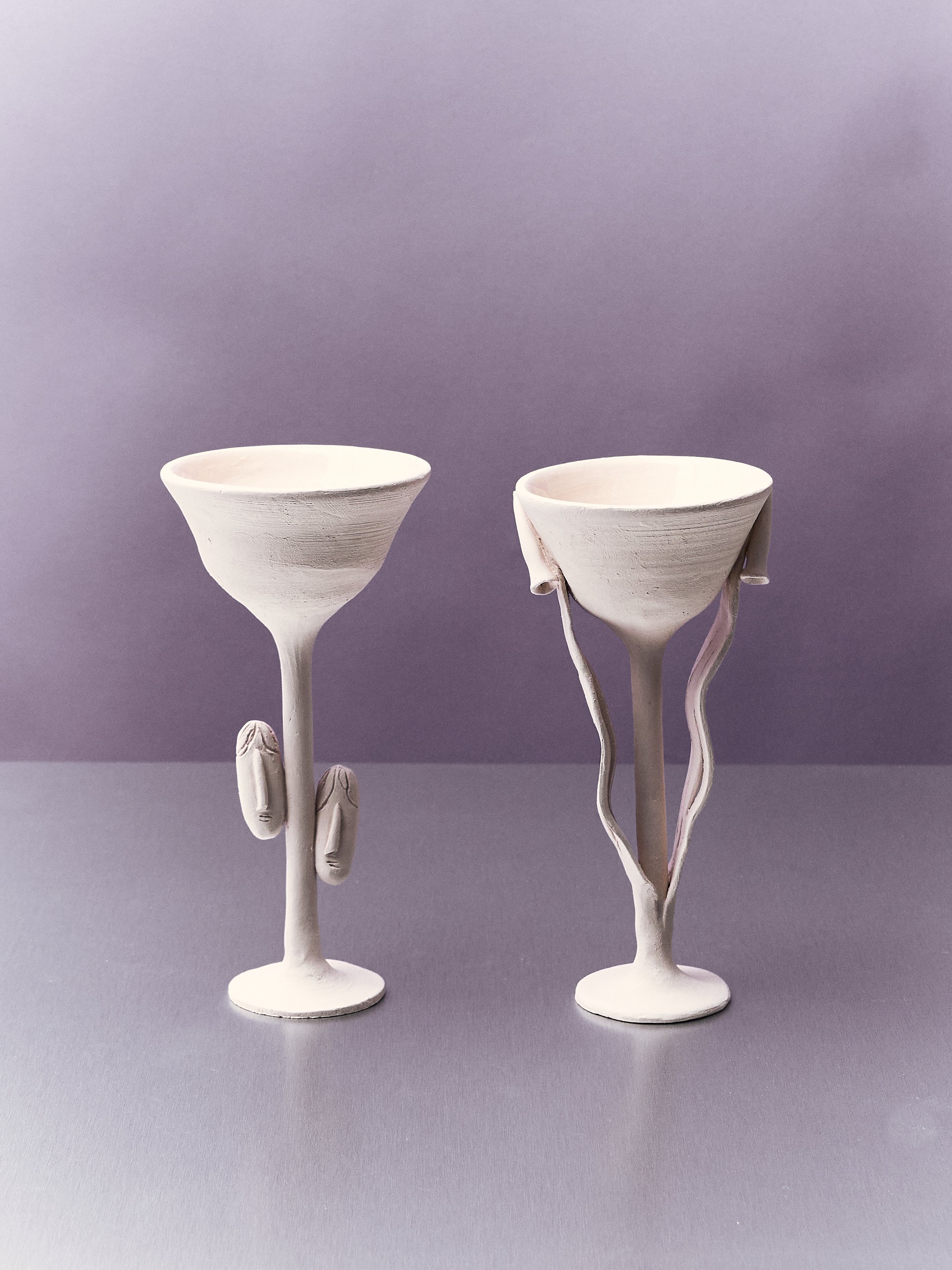 Due bicchieri artistici bianchi con forme umane, uno con facce e mani modellate, l'altro con linee ondulate sul gambo, su sfondo viola.