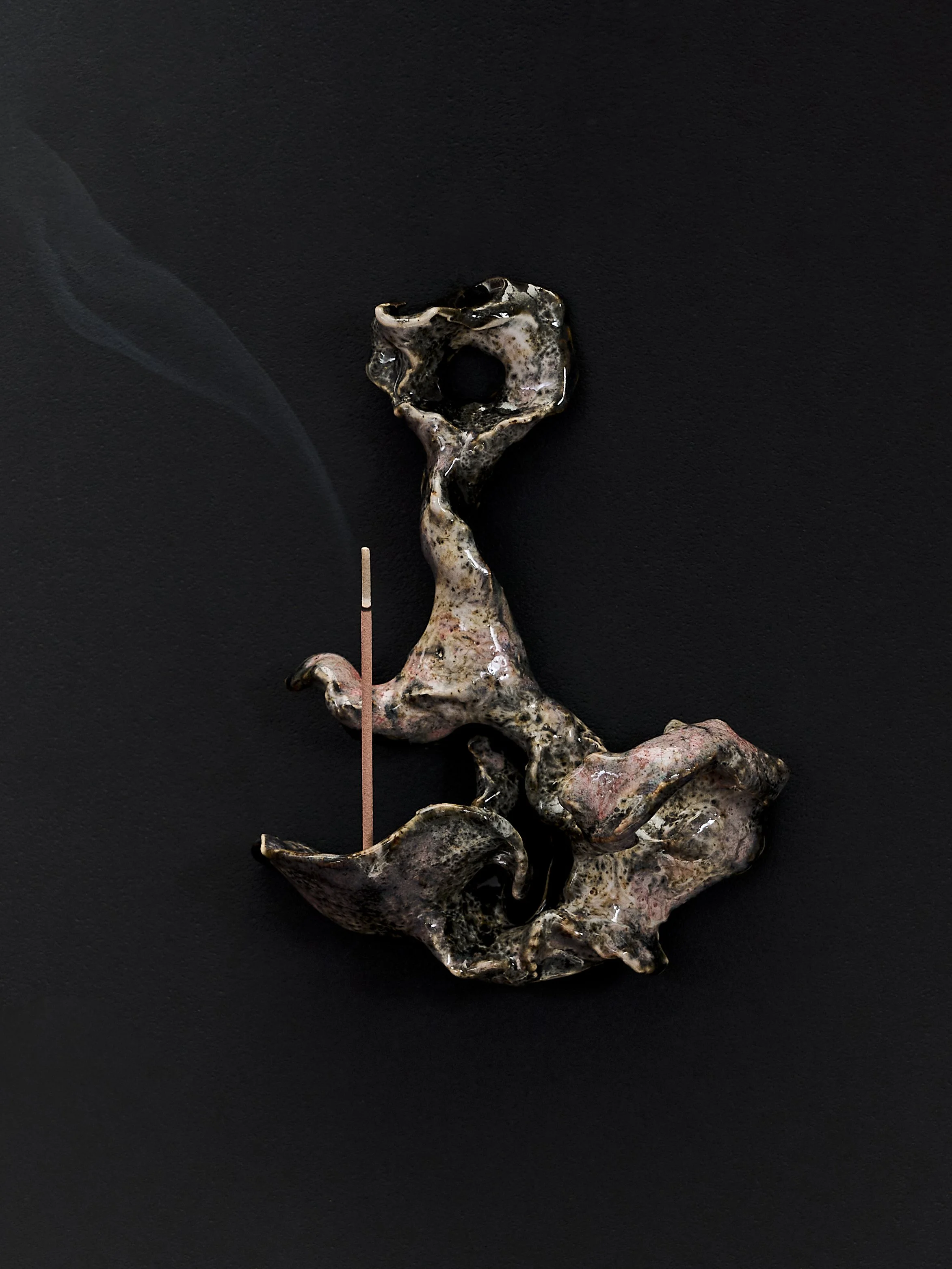 Oggetto astratto che ricorda la forma di una lettera 'J' su sfondo nero, con un bastoncino di incenso accanto.