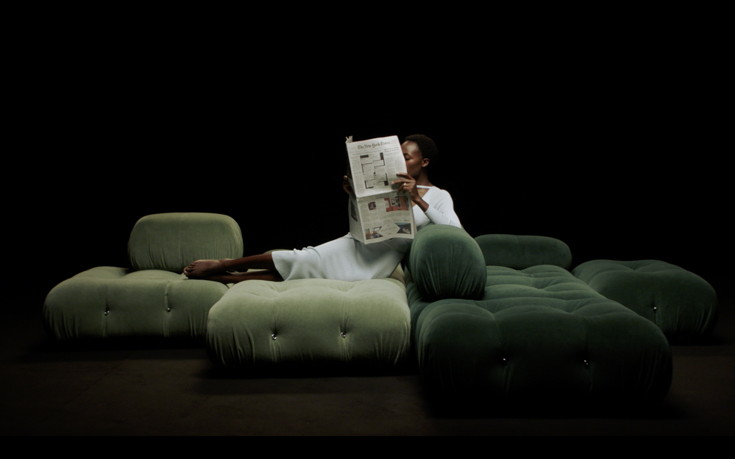 Una persona con pelle scura e vestiti bianchi distesa su un divano verde scuro, che legge un giornale in un ambiente buio.